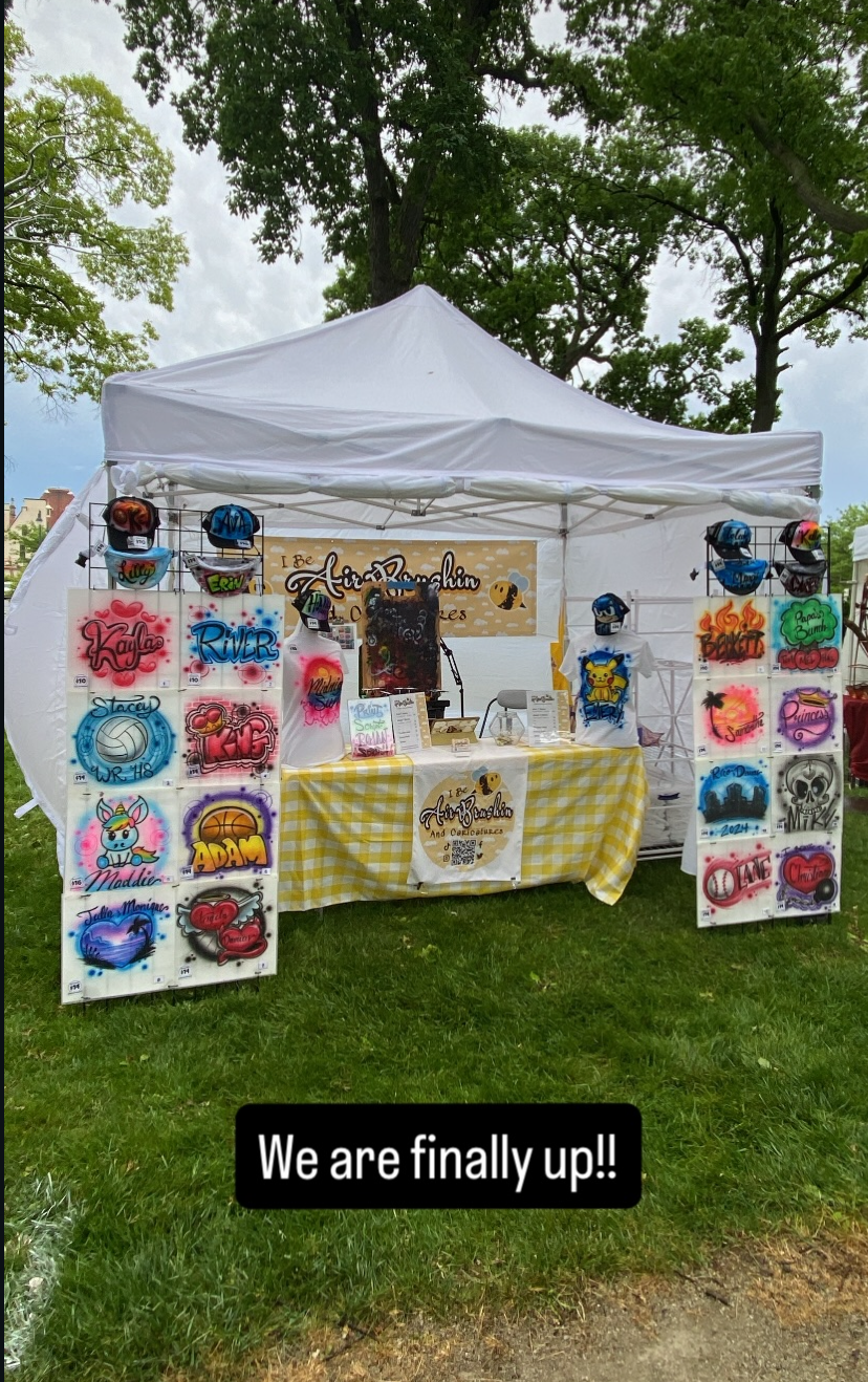 A white outdoor tent with a yellow and white checkered tablecloth displaying custom painted helmets, colorful painted signs with names and designs, and a sign saying 'I Be AirBrushin and Caricatures.' The scene is set in a park with green trees and a cloudy sky in the background.