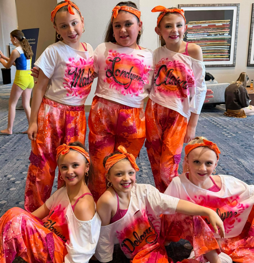 Five young girls in colorful neon costumes, with orange bandanas on their heads, posing together at an indoor event.