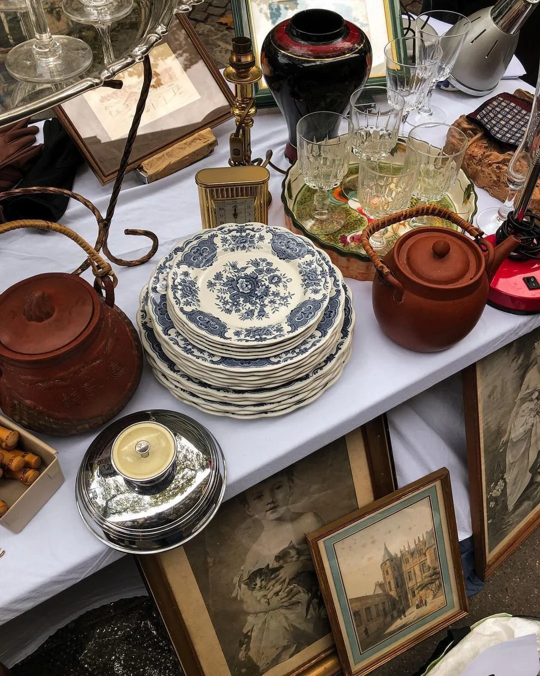 Une table avec de la vaisselle en porcelaine à motifs bleus, deux théières en terre cuite, plusieurs verres à vin, un cadre avec une photo de jeune fille, des peintures encadrées, et divers objets décoratifs.