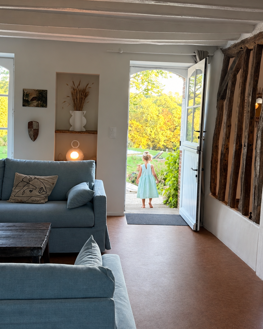 Une jeune fille avec une robe bleue claire entre dans un jardin par une porte entrouverte, laissant apparaître un espace extérieur verdoyant ensoleillé.