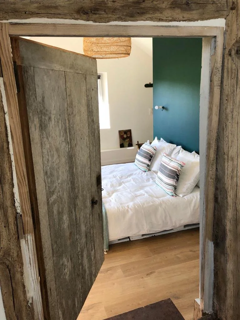 Vue d'une chambre à coucher avec un lit blanc, des oreillers à motifs, un mur vert, une fenêtre et un plafond avec un luminaire en osier, vue partielle à travers une porte en bois brut.