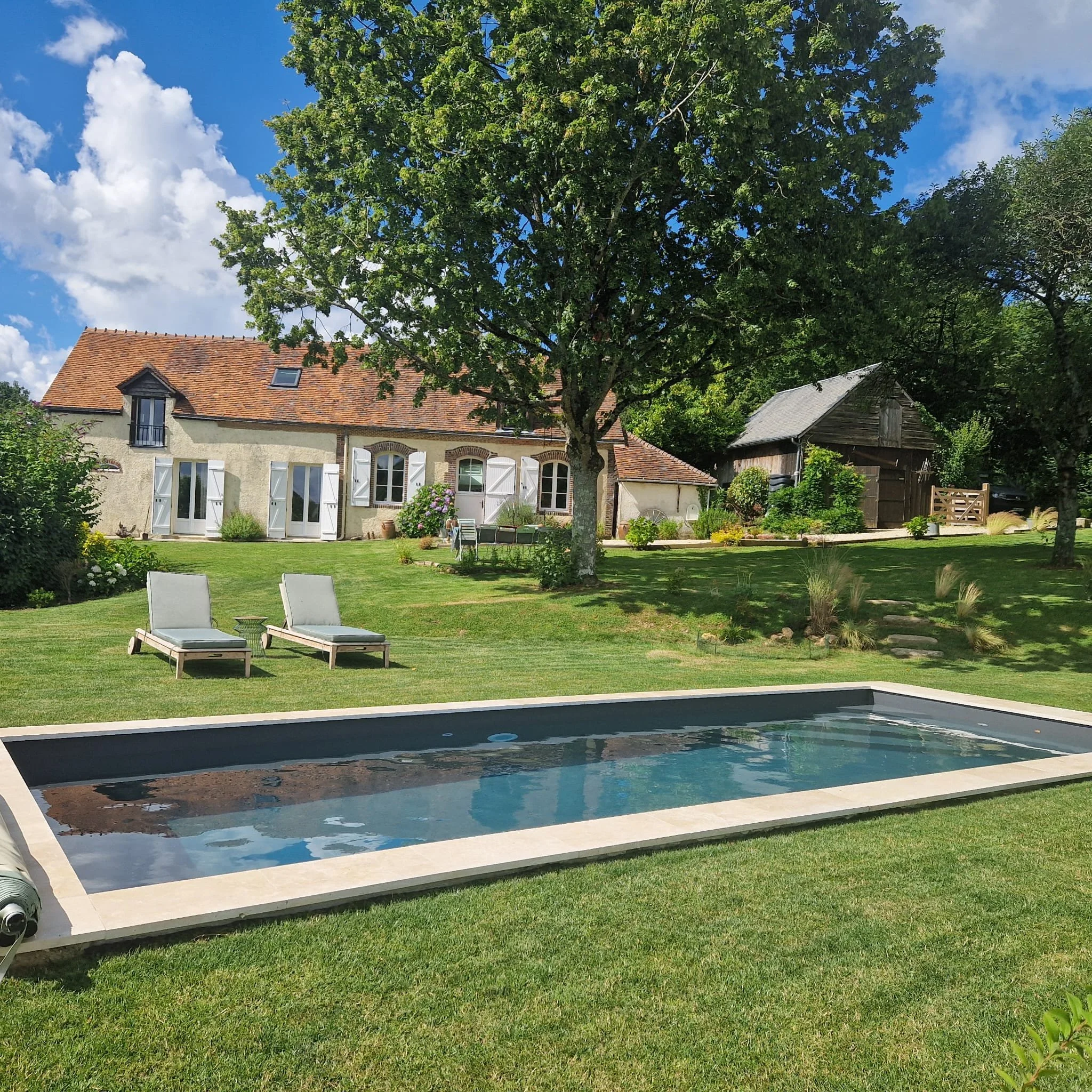 Une maison avec un toit en tuiles rouges, entourée d'un jardin verdoyant avec des meubles et une piscine, sous un ciel bleu avec quelques nuages.