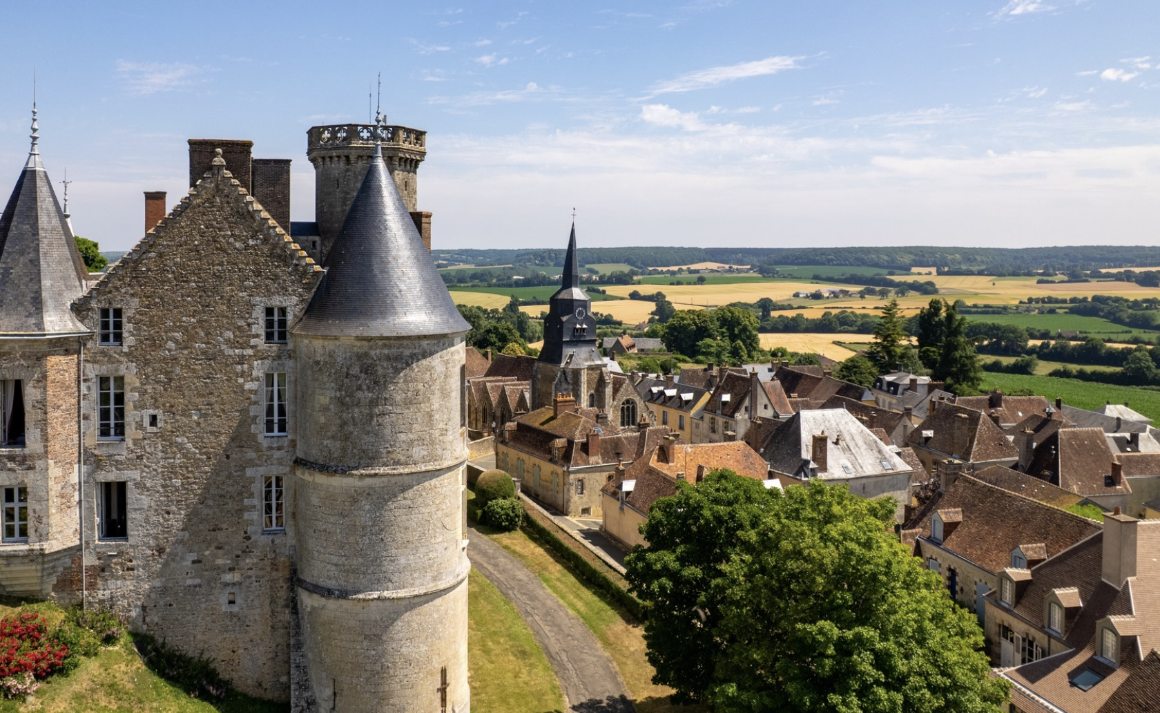 Un village médiéval avec des maisons en pierre et des tours, entouré de champs agricoles verts et dorés, sous un ciel en partie nuageux.