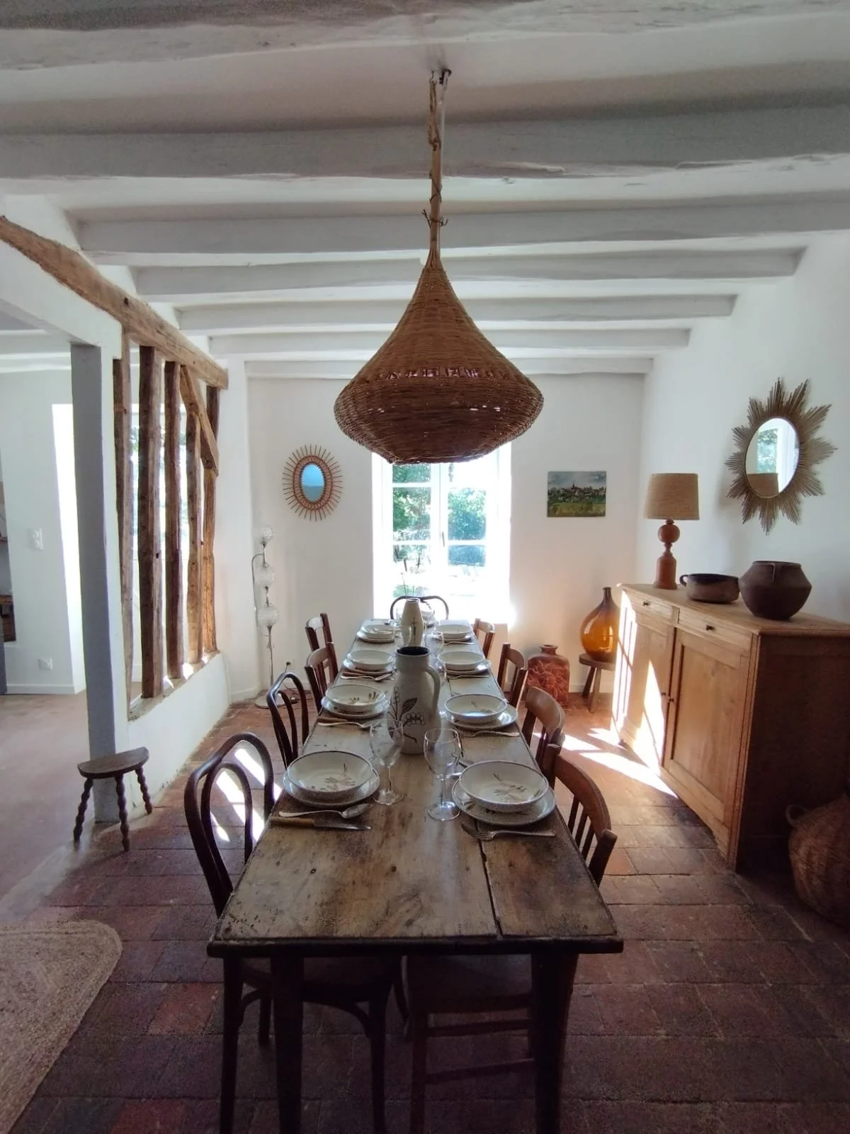 Salle à manger rustique avec une grosse lampe en osier suspendue, table en bois avec assiettes, verres et carafes, fenêtres apportant de la lumière naturelle, meuble en bois avec des décorations