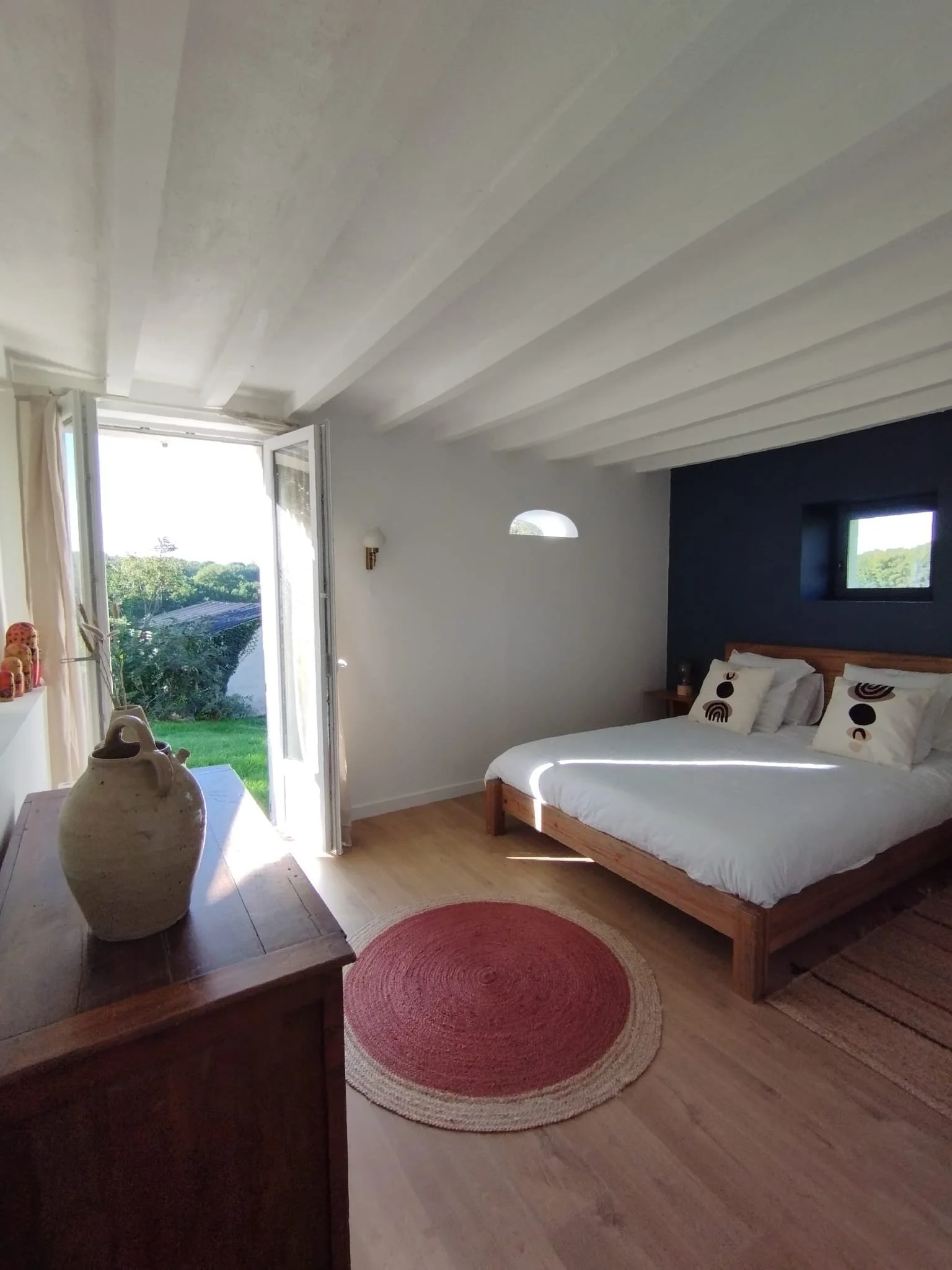 Chambre avec lit en bois, oreillers à motifs, petite fenêtre, porte-fenêtre grande laissant entrer la lumière, tapis en forme de cercle rouge-orange, décoration simple avec pot en céramique et vue sur la nature. 