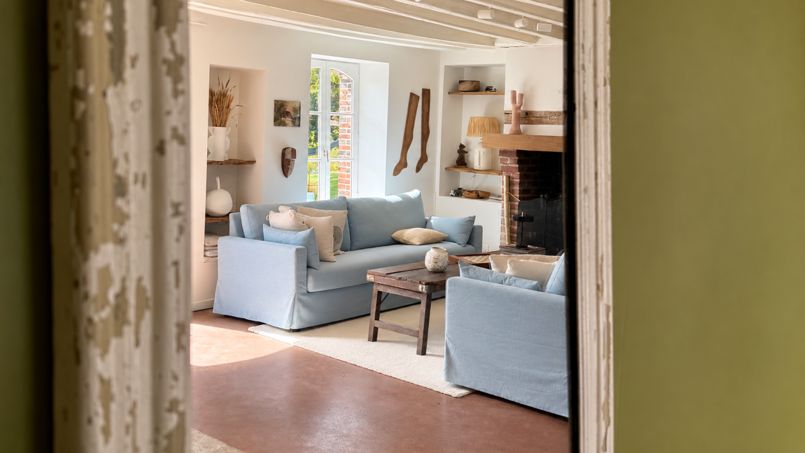 Salon avec deux canapés bleus, cheminée en brique, étagères en bois, décoration rustique et fenêtre donnant sur un jardin.