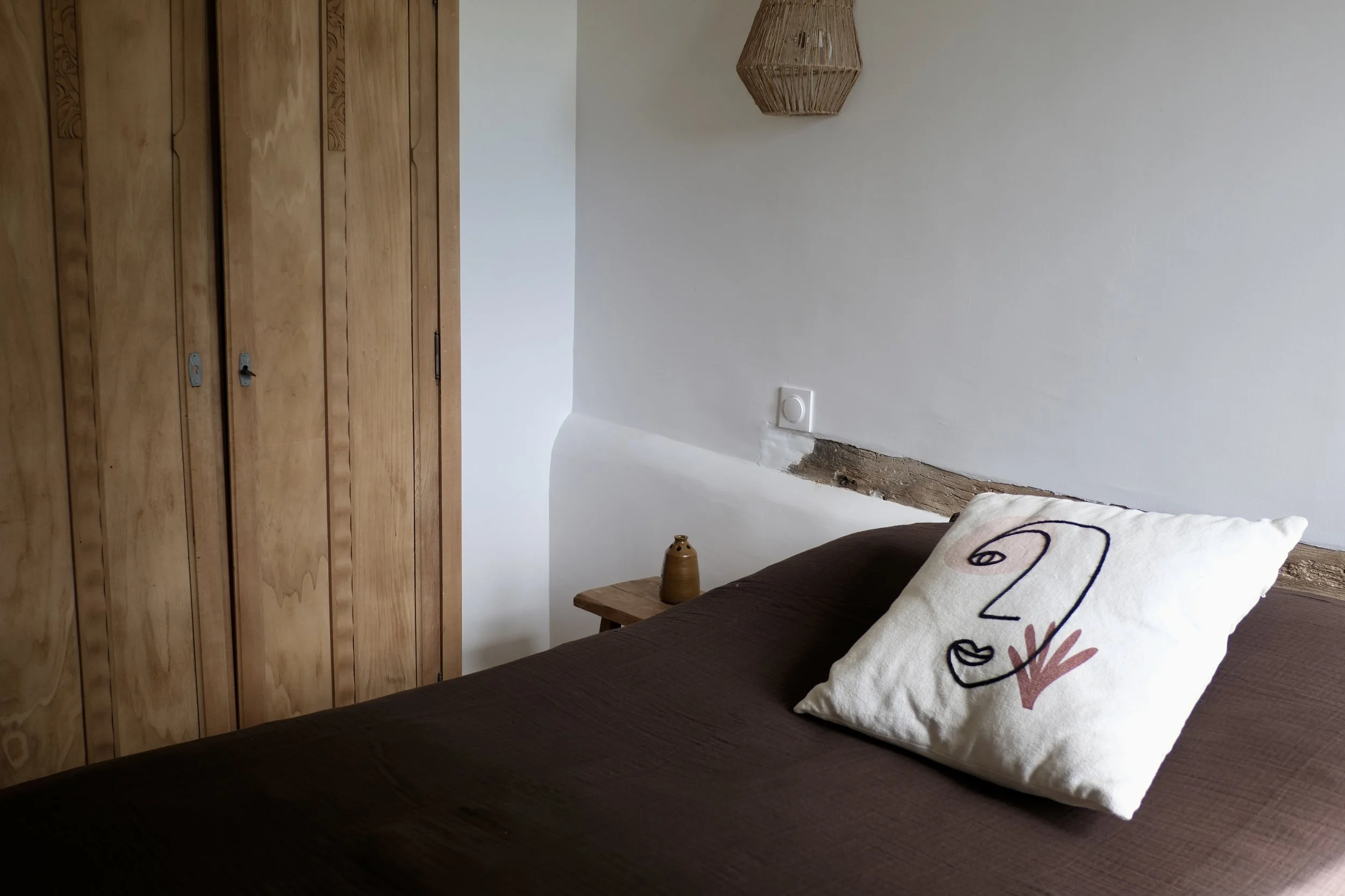 Une chambre avec un mur blanc, une armoire en bois, un lit avec une couverture marron et un coussin décoratif blanc avec un dessin noir et rouge, une petite table en bois avec une petite jarre en terre cuite, et une lampe suspendue en rotin.