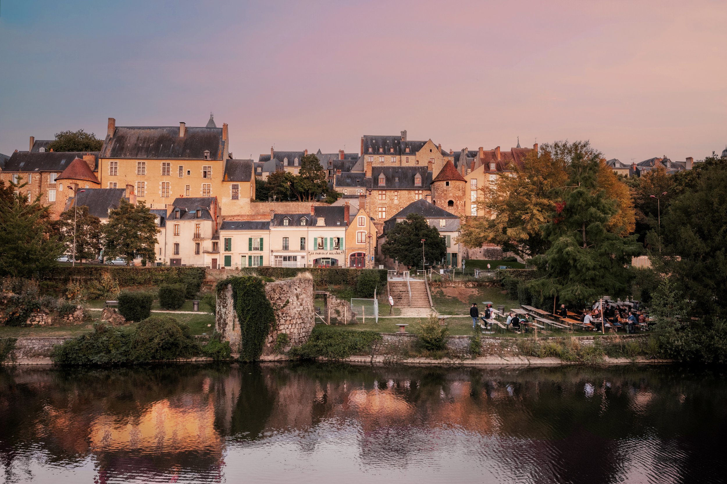 Vue d'un ancien village avec des bâtiments en pierre et toiture en ardoise, entouré d'arbres, avec une rivière au premier plan et un ciel au crépuscule.