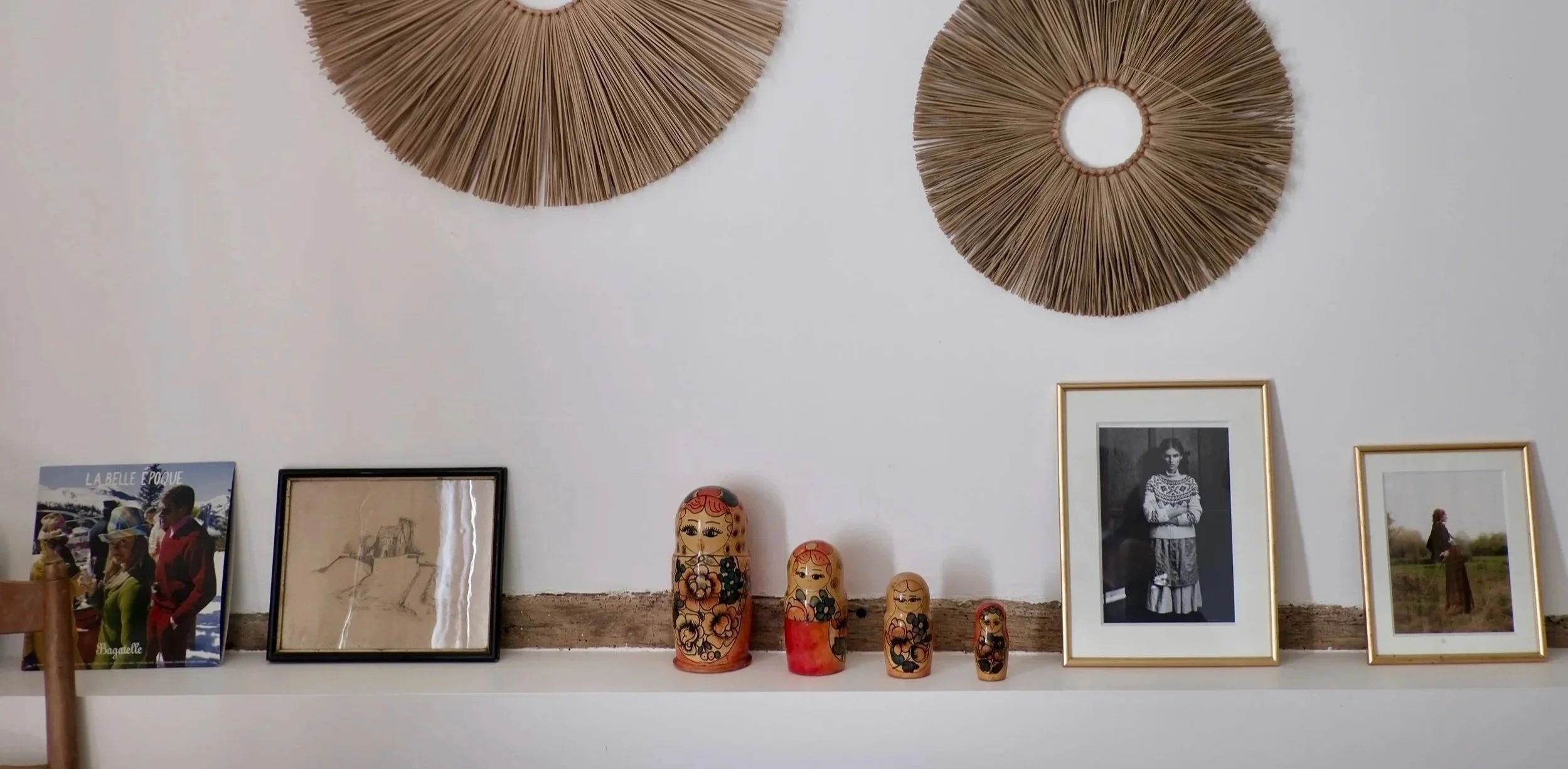 Décor mural avec deux grands ronds en bois ou paille, plusieurs cadres avec photos et illustrations, et des poupées russes colorées en céramique, dans un intérieur élégant.
