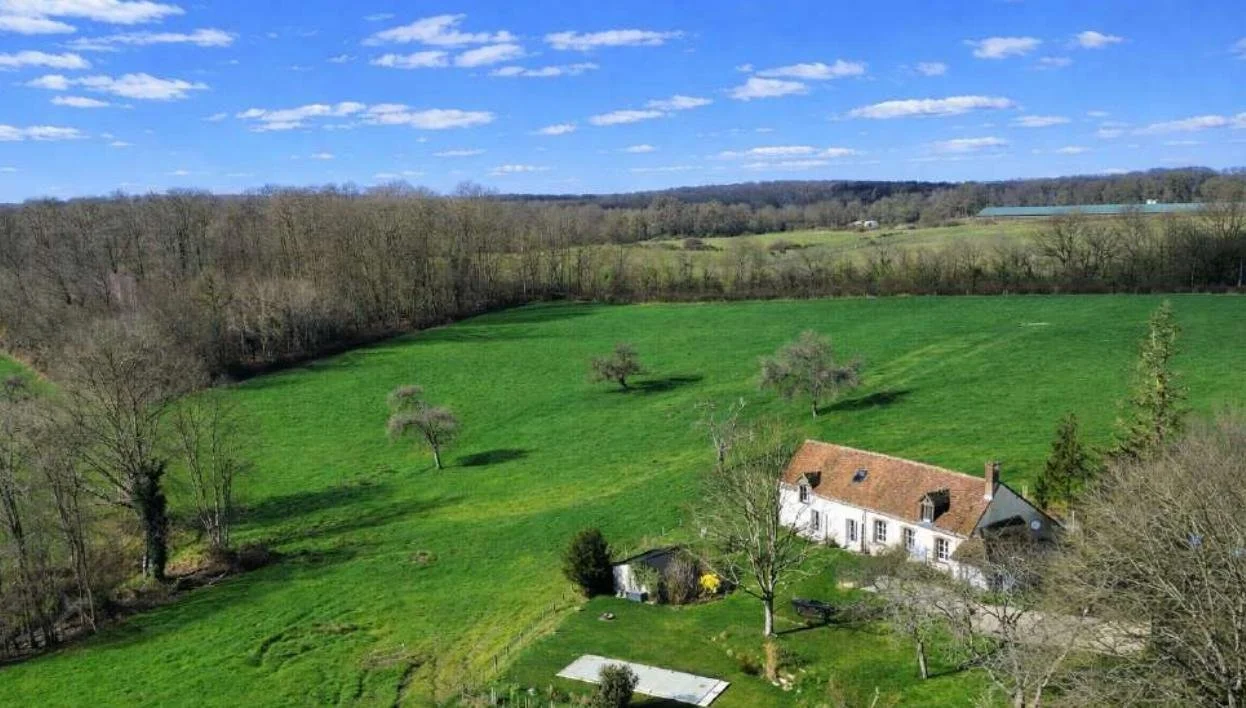 Paysage rural avec une maison en pierre, un grand terrain vert, des arbres et un ciel bleu avec quelques nuages.