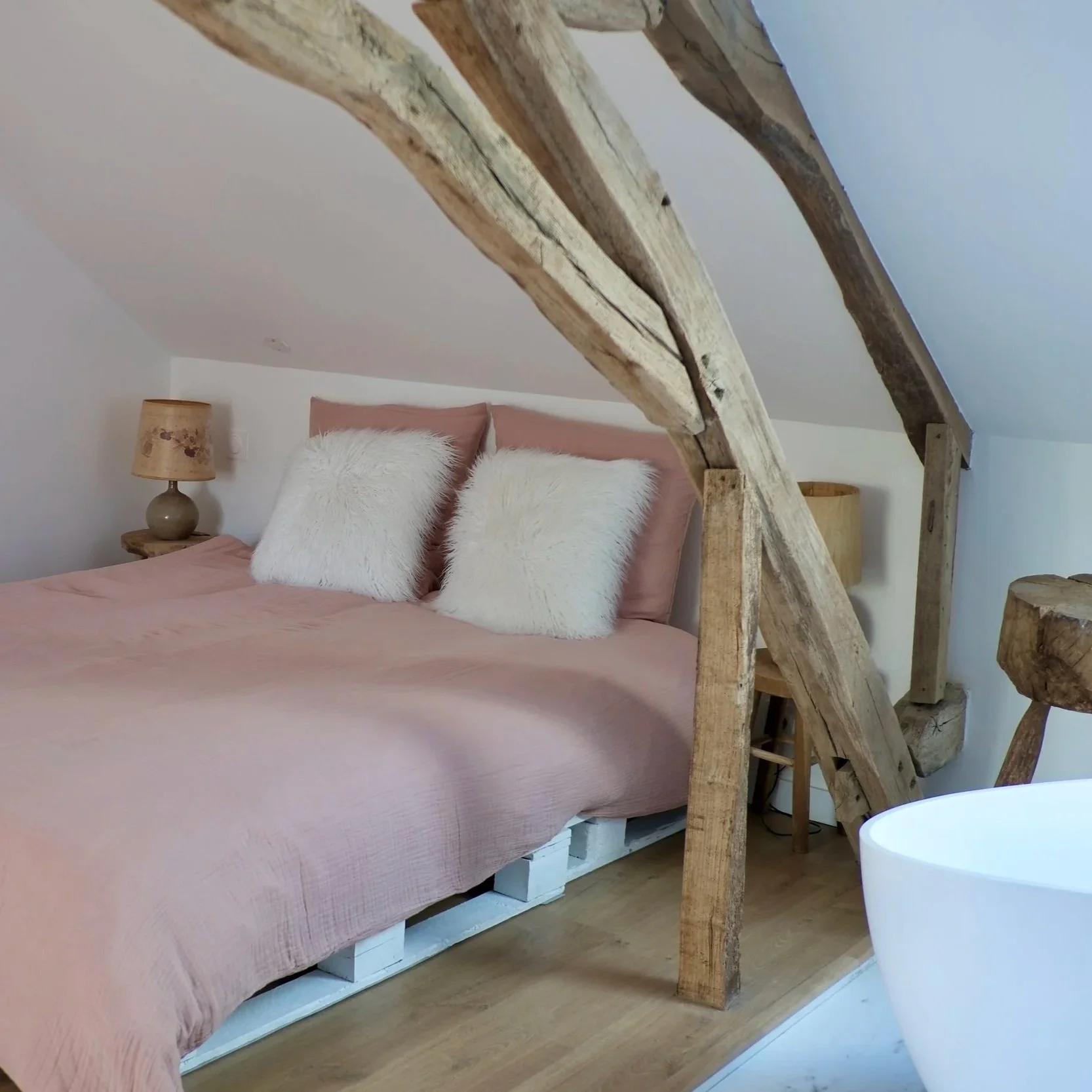 Un lit dans une chambre avec une tête de lit rose, deux oreillers blancs en fourrure, une lampe de chevet en céramique et une pièce en bois brut dans un intérieur au plafond en pente.