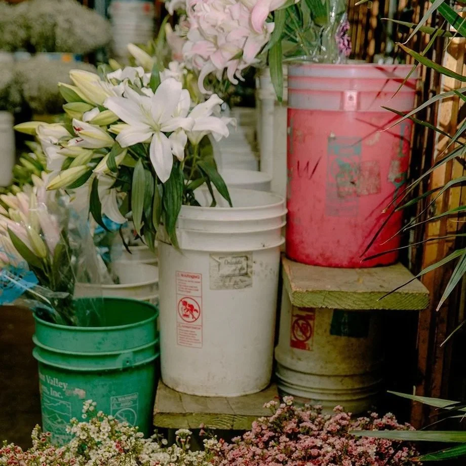 Pots de fleurs avec des lys blancs et des fleurs roses, disposés sur un support en bois dans une jardinerie ou un espace de fleurs.