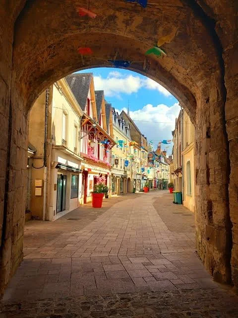Vue d'une rue piétonne encadrée par une arche en pierre, avec bâtiments colorés et un ciel bleu avec des nuages.