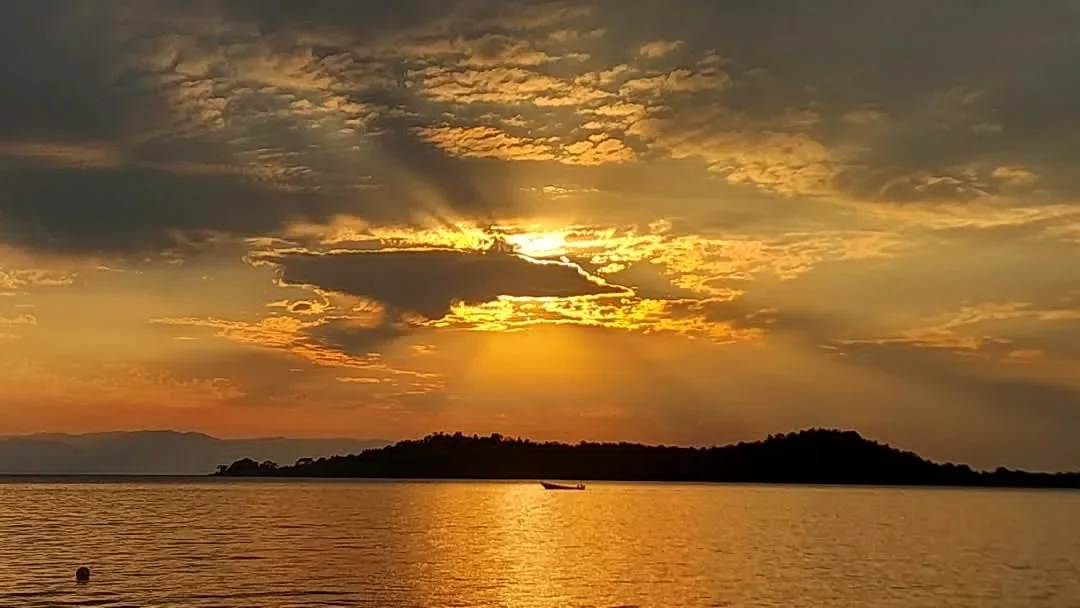 Golden sunset over Lake Tanganyika, Tanzania, with dramatic clouds, sun rays reflecting on the water and a small boat silhouetted against the island.