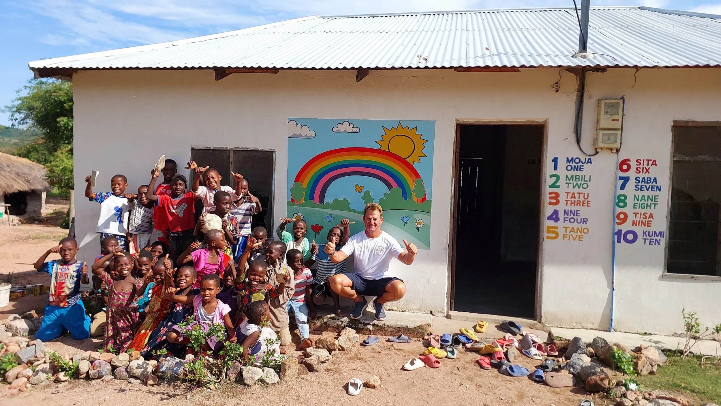 Group of smiling children and a visitor outside a colourful community learning centre near Kipili, Tanzania, illustrating local village life and education near Lake Shore Lodge.