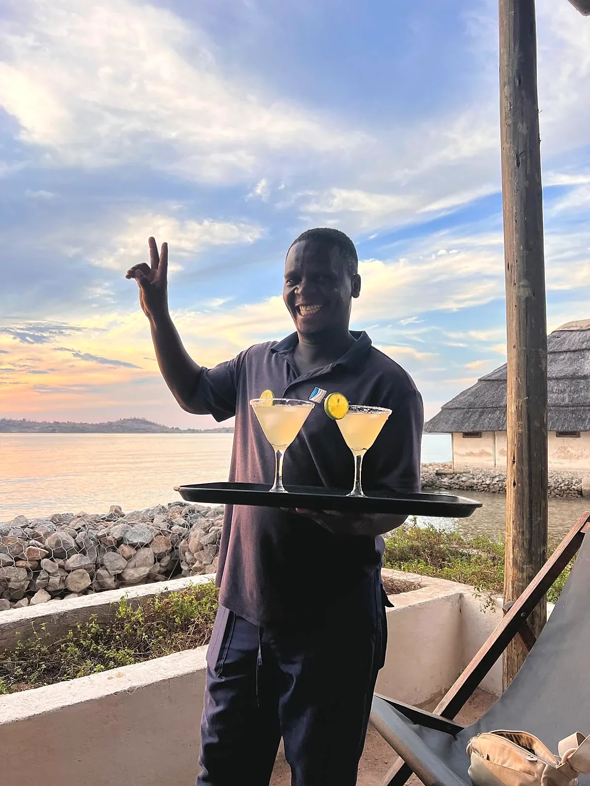Smiling Lake Shore Lodge team member serving two cocktails overlooking Lake Tanganyika at sunset in Kipili, Tanzania.