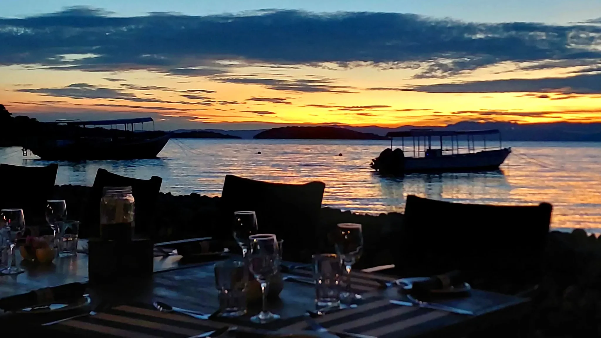 Waterfront dinner tables set for evening dining at Lake Shore Lodge, overlooking boats silhouetted against a golden Lake Tanganyika sunset in Tanzania.