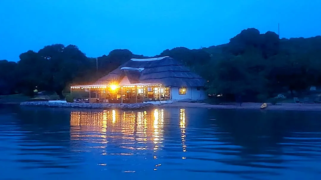 Thatched-roof lakeside restaurant glowing at dusk, reflected in calm blue water