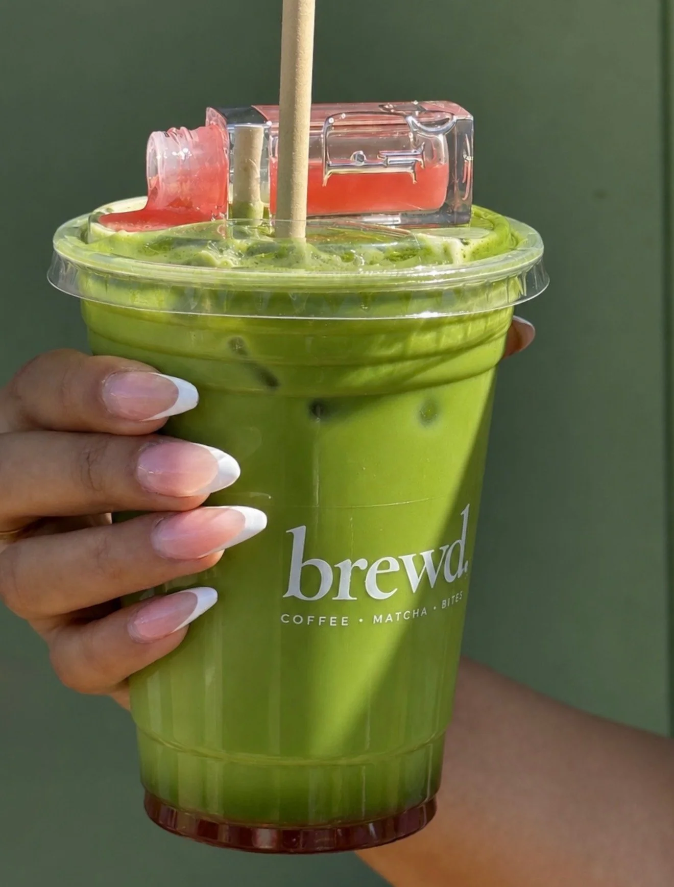 Close-up of a person holding a green iced matcha latte with a plastic lid, showing a red and clear plastic object on top of the drink.