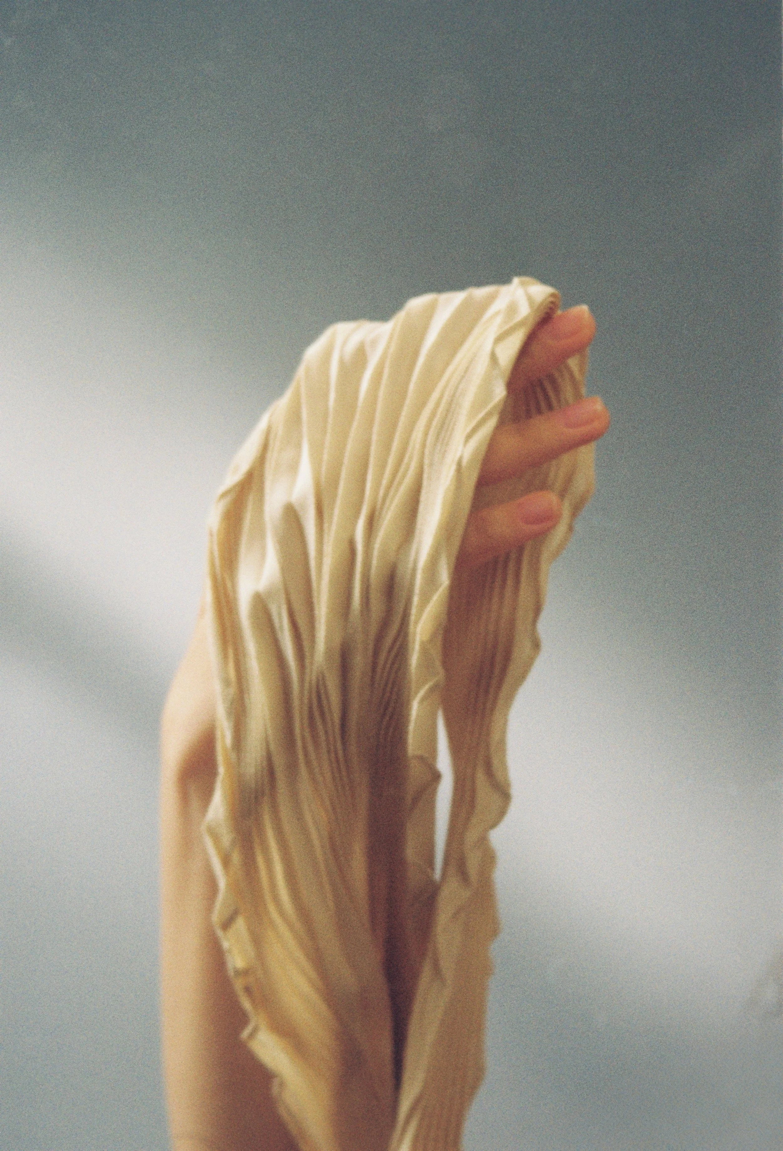 Hand holding a pleated cream scarf against a grey background.