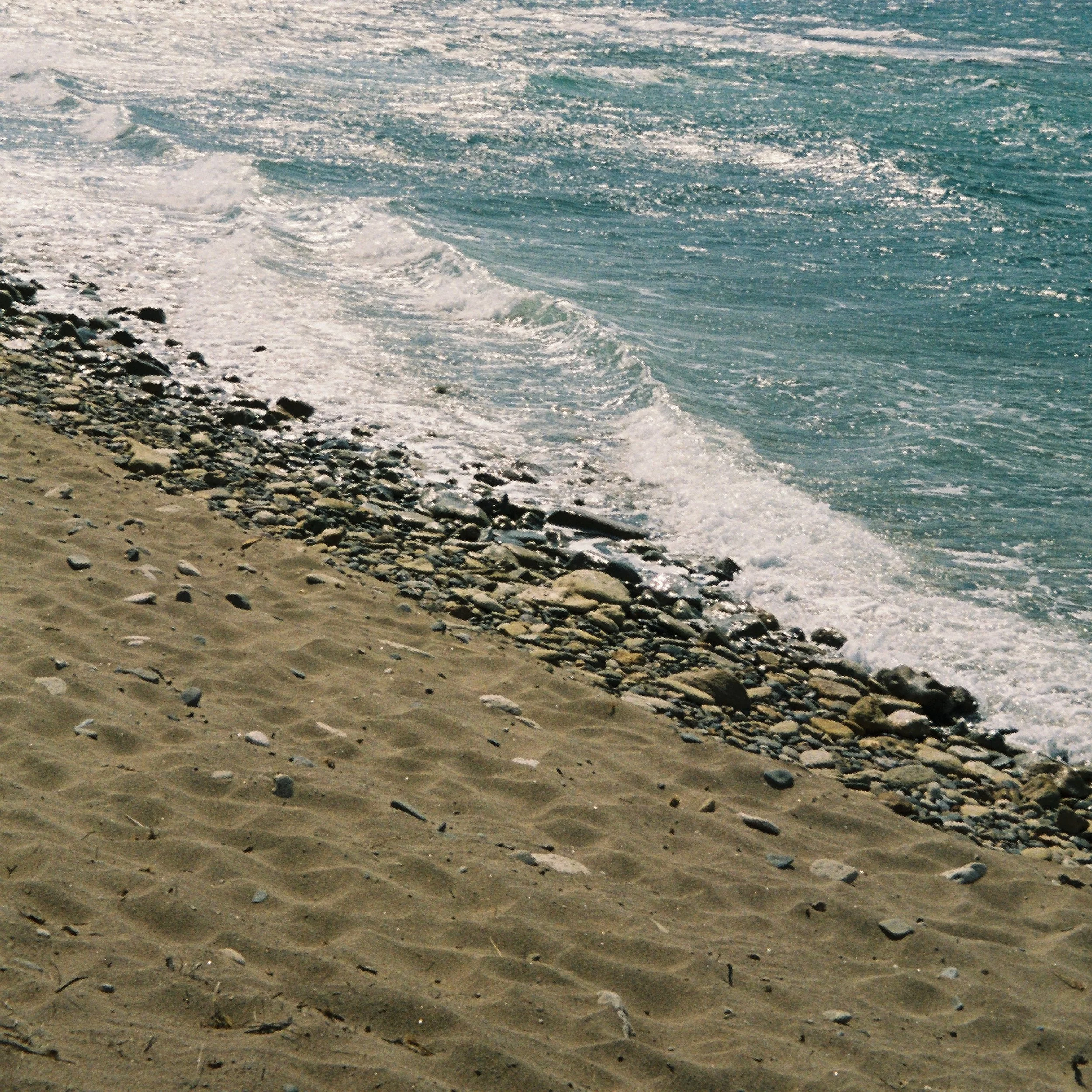 A sandy beach with small rocks and pebbles, and ocean waves gently lapping against the shore.