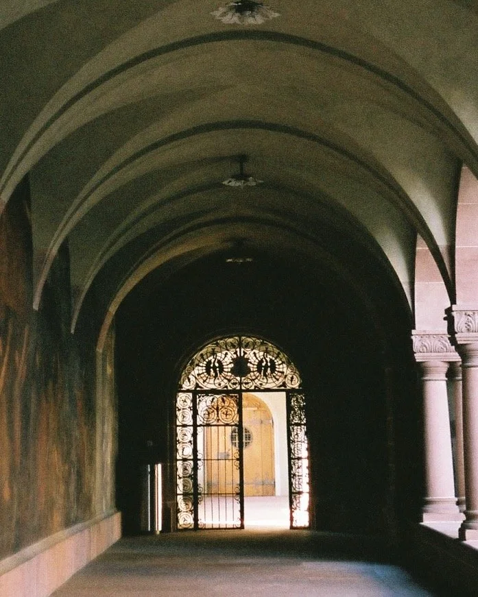 A dark arched hallway with ornamental wrought-iron gate and stone columns on the right side, leading outside to a bright courtyard with yellow walls.