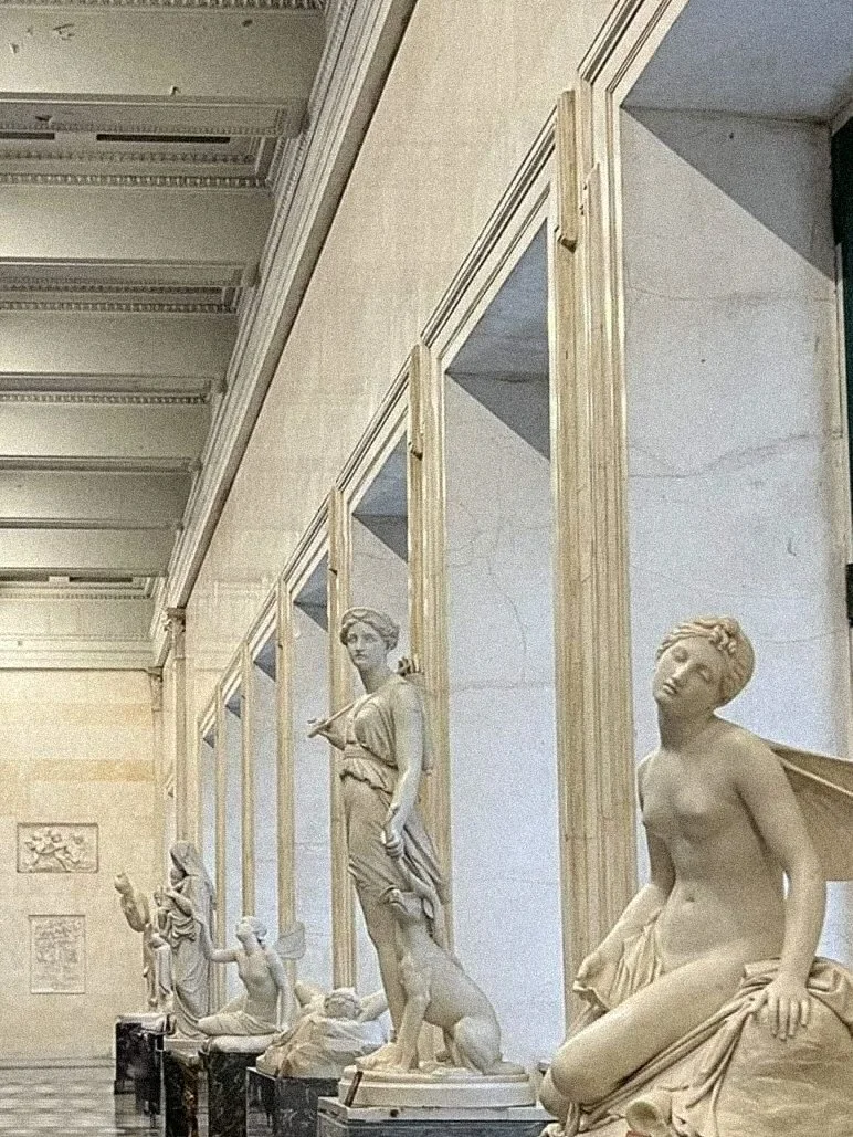 A row of classical marble sculptures displayed in Hermitage corridor, with some sculptures framed in wooden moldings, along a marble wall .