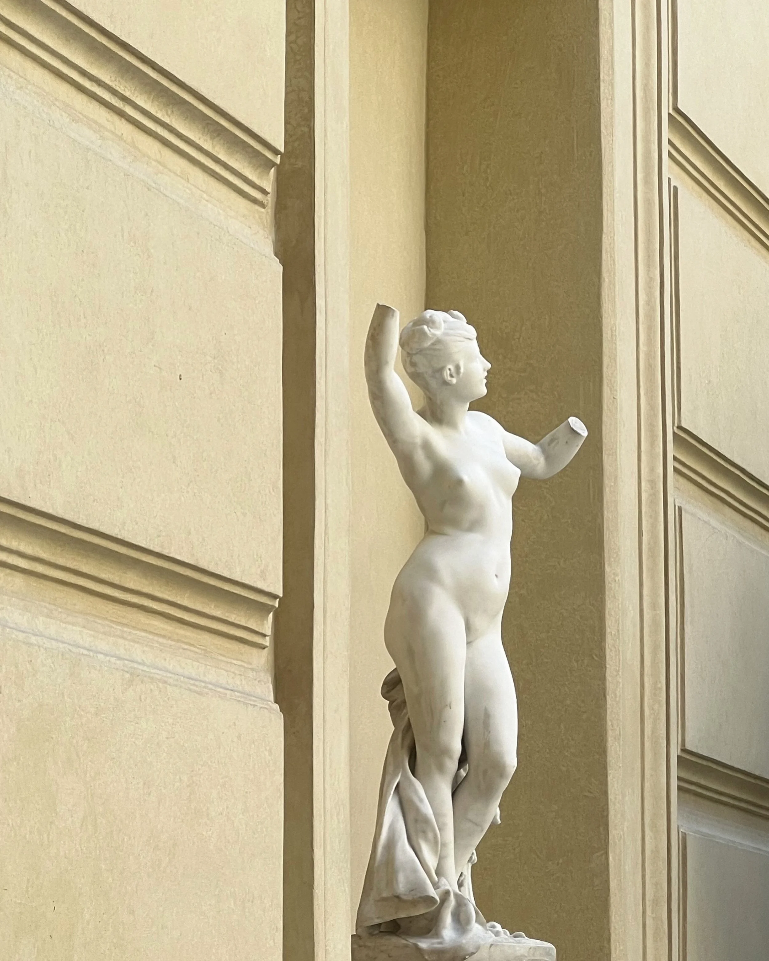 A white marble sculpture of a woman with raised arms, standing against a beige wall with decorative molding.