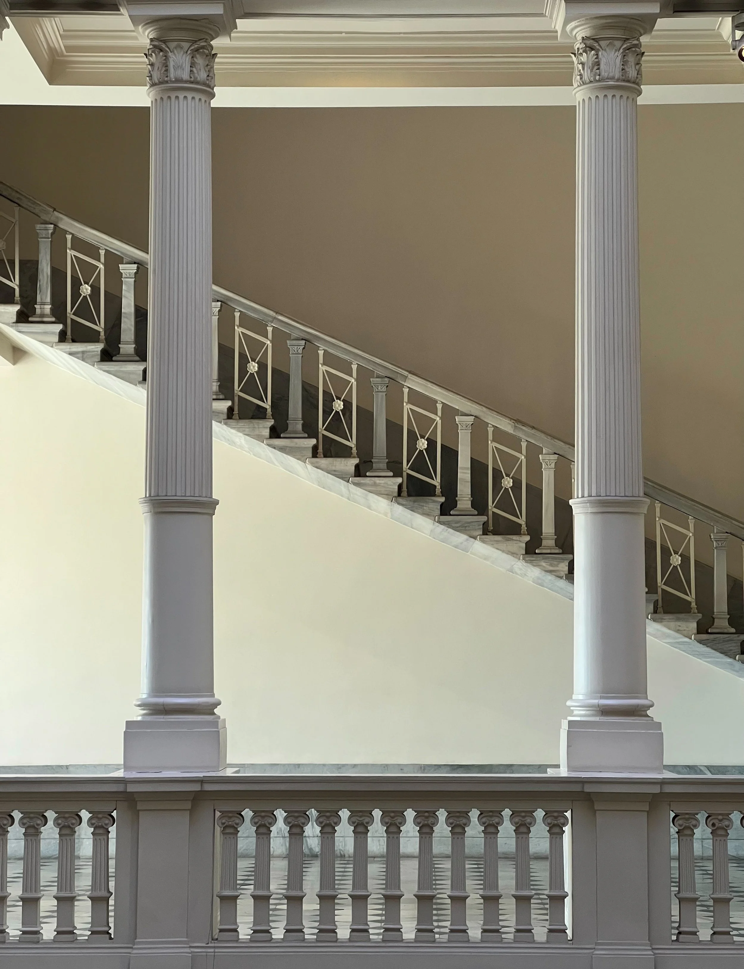 White balcony with large decorative columns and ornate railings, overlooking a marble staircase in a building interior.