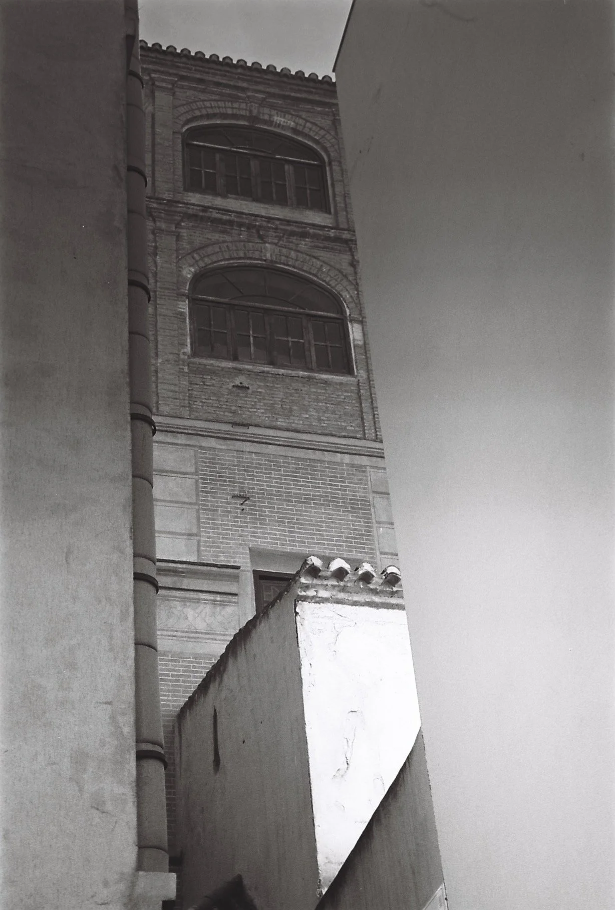 Black and white photo of a tall brick building viewed from a narrow alleyway between two walls, with windows, a tiled roof, and a pipe running down the side of the building.