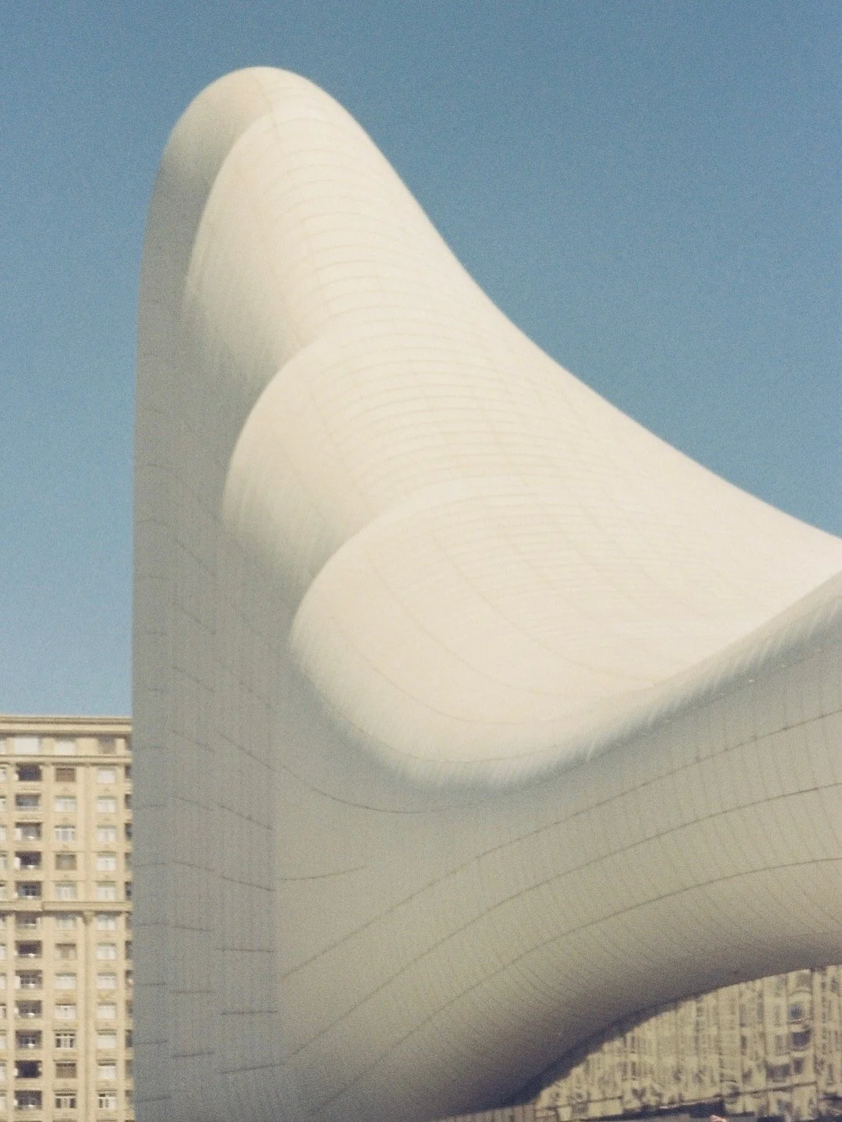The image shows a section of a modern, curved architectural building with a white exterior, set against a clear blue sky, with a tall residential building in the background.