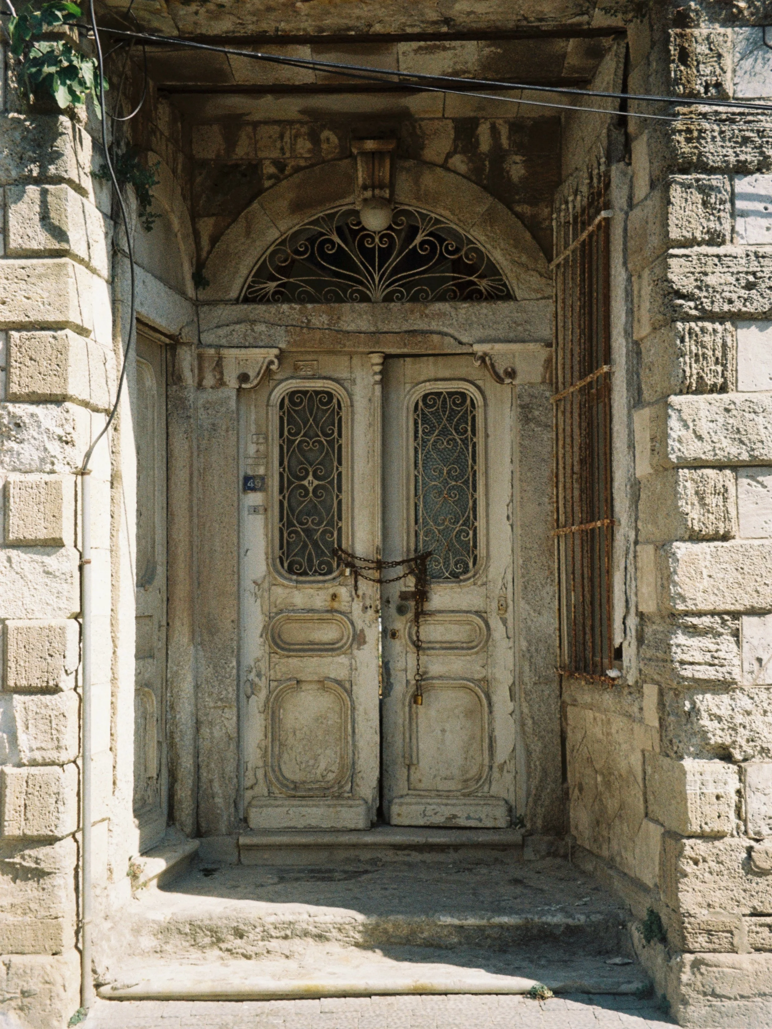 An old stone doorway with ornate wrought iron detailing on the double doors, secured with a rusted chain and padlock, and a stone archway above.