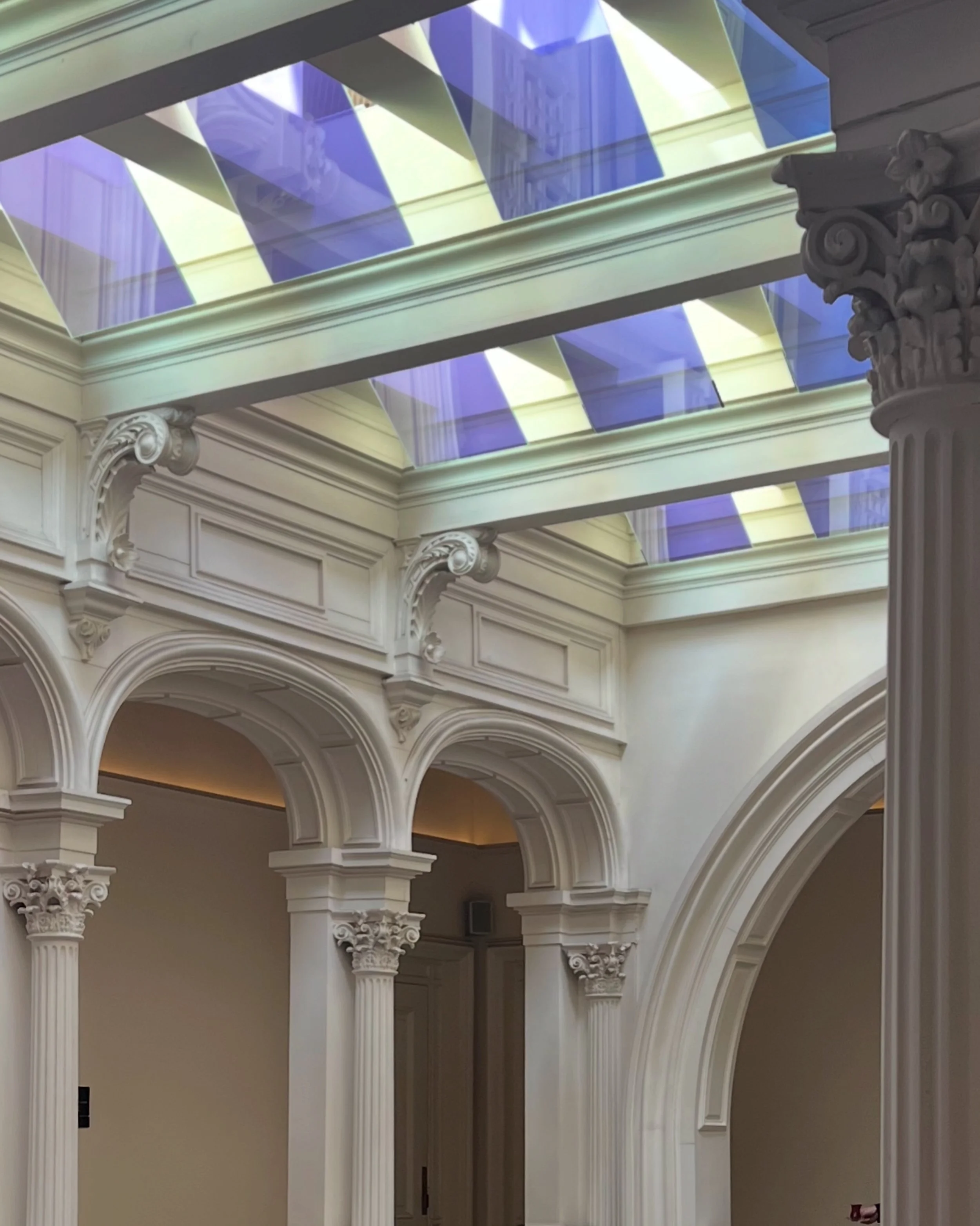 Interior of a building with classical architectural features, including tall white Corinthian columns with ornate capitals, decorative archways, and a decorative ceiling with large skylights.