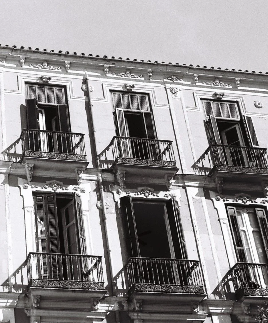 Gray building with multiple open windows and balconies with decorative wrought iron railings, shadows cast by the balconies on the facade.
