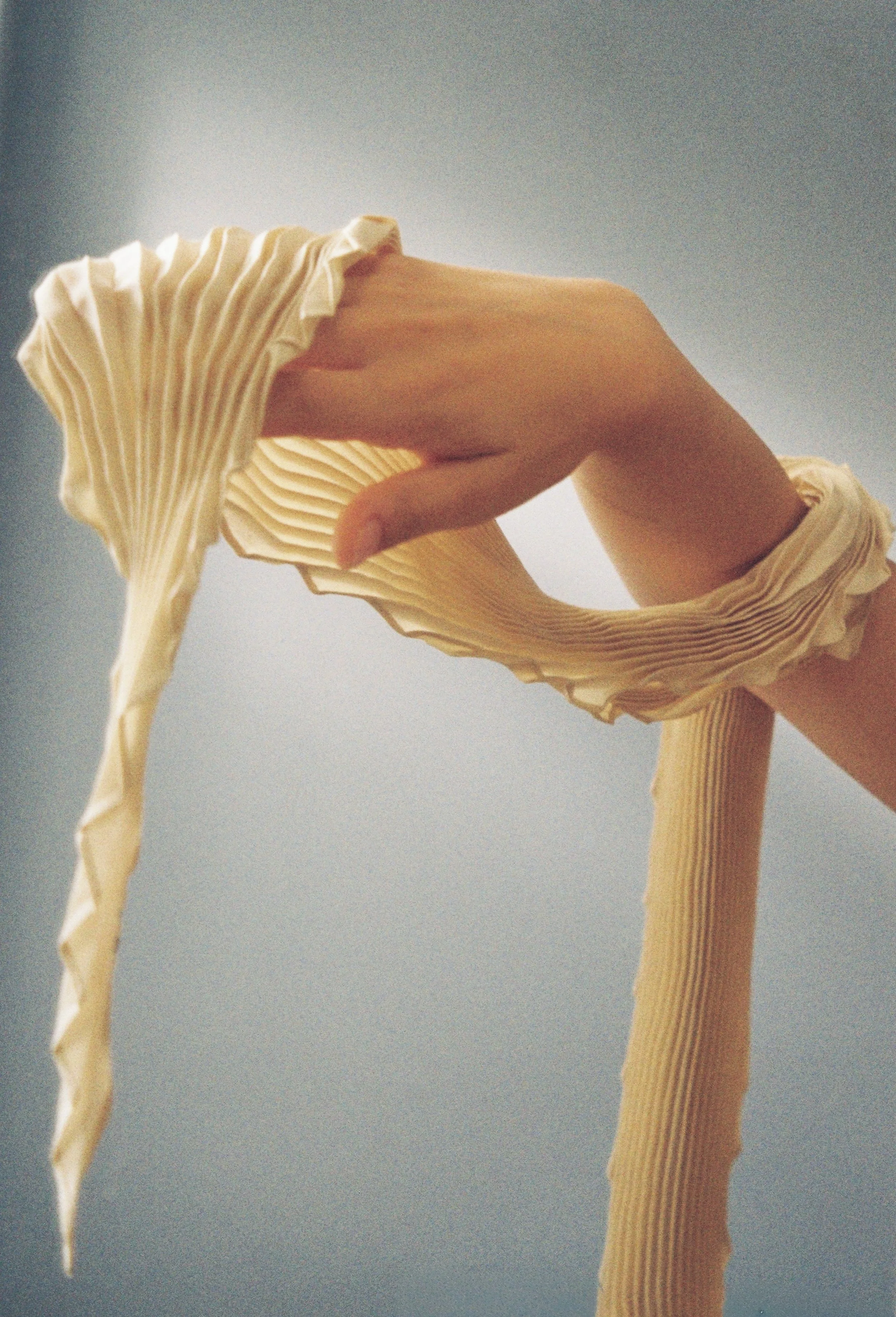 Close-up of a hand holding a pleated, cream-colored scarf against a grey background.