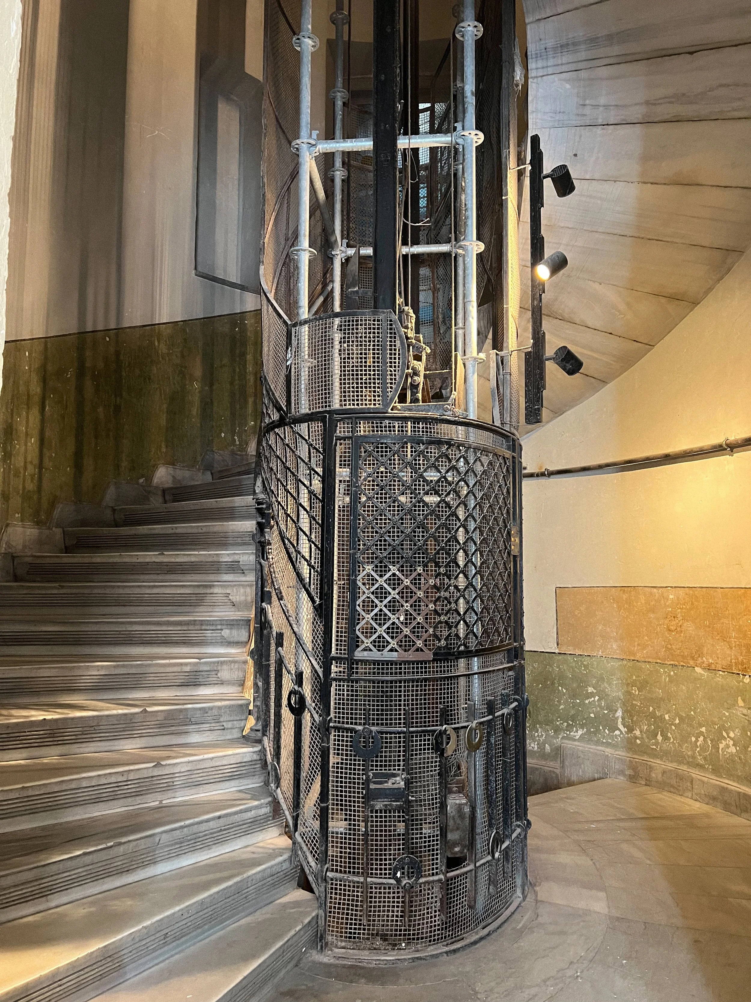 An old metal elevator with a cylindrical cage structure located at the bottom of stairs in a concrete building.