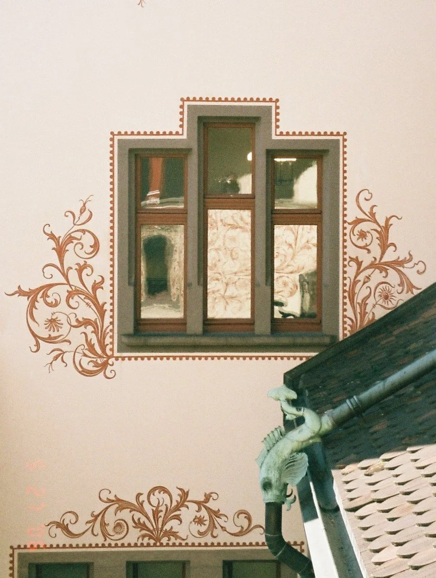 A close-up of a roof with a decorative dragon-shaped gutter, a window with a wooden frame, and ornate floral patterns painted around the window on a building wall.