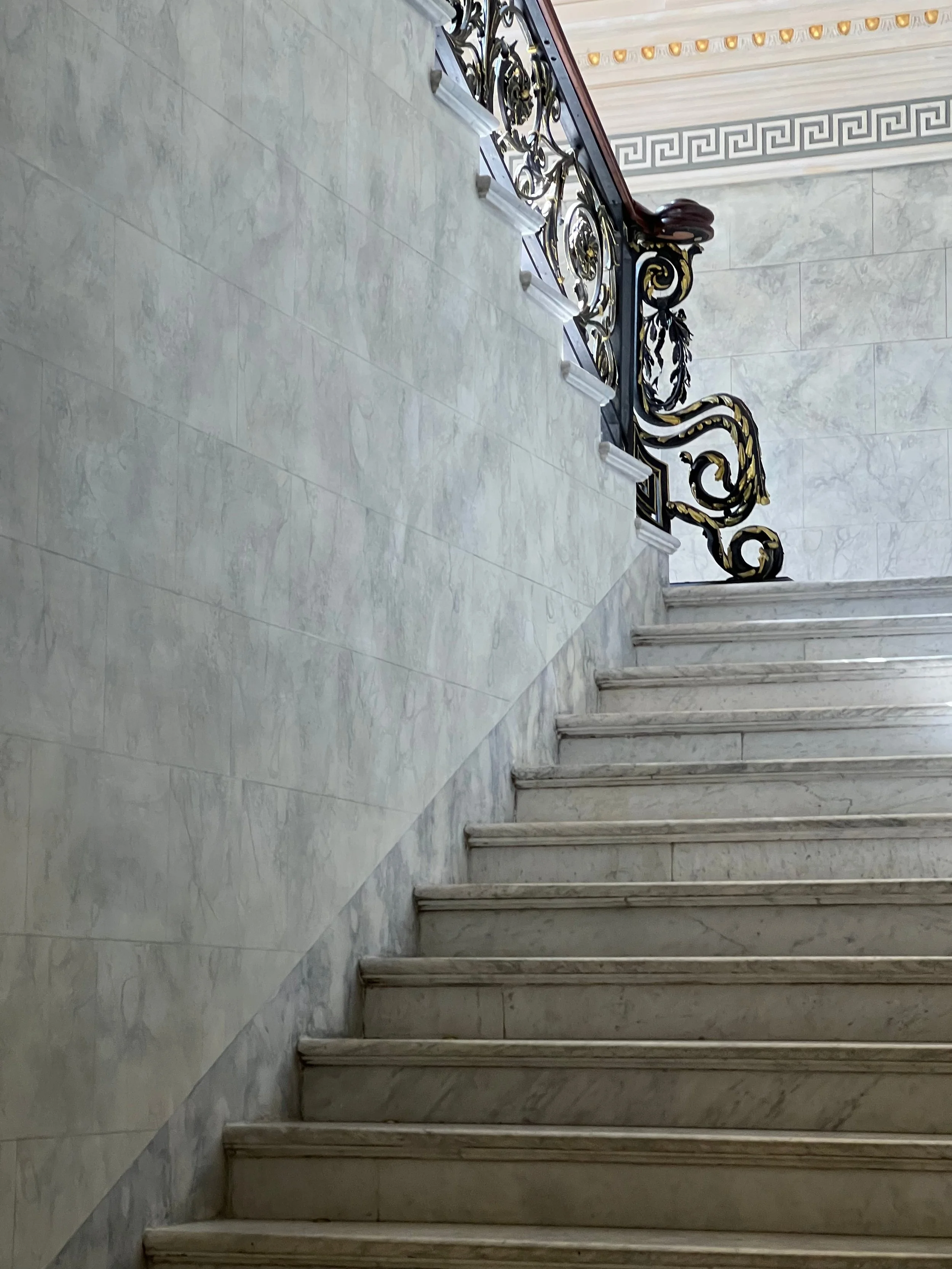 Decorative staircase with ornate black and gold wrought iron railing and marble steps in a classical interior with Greek key patterned border and marble walls.