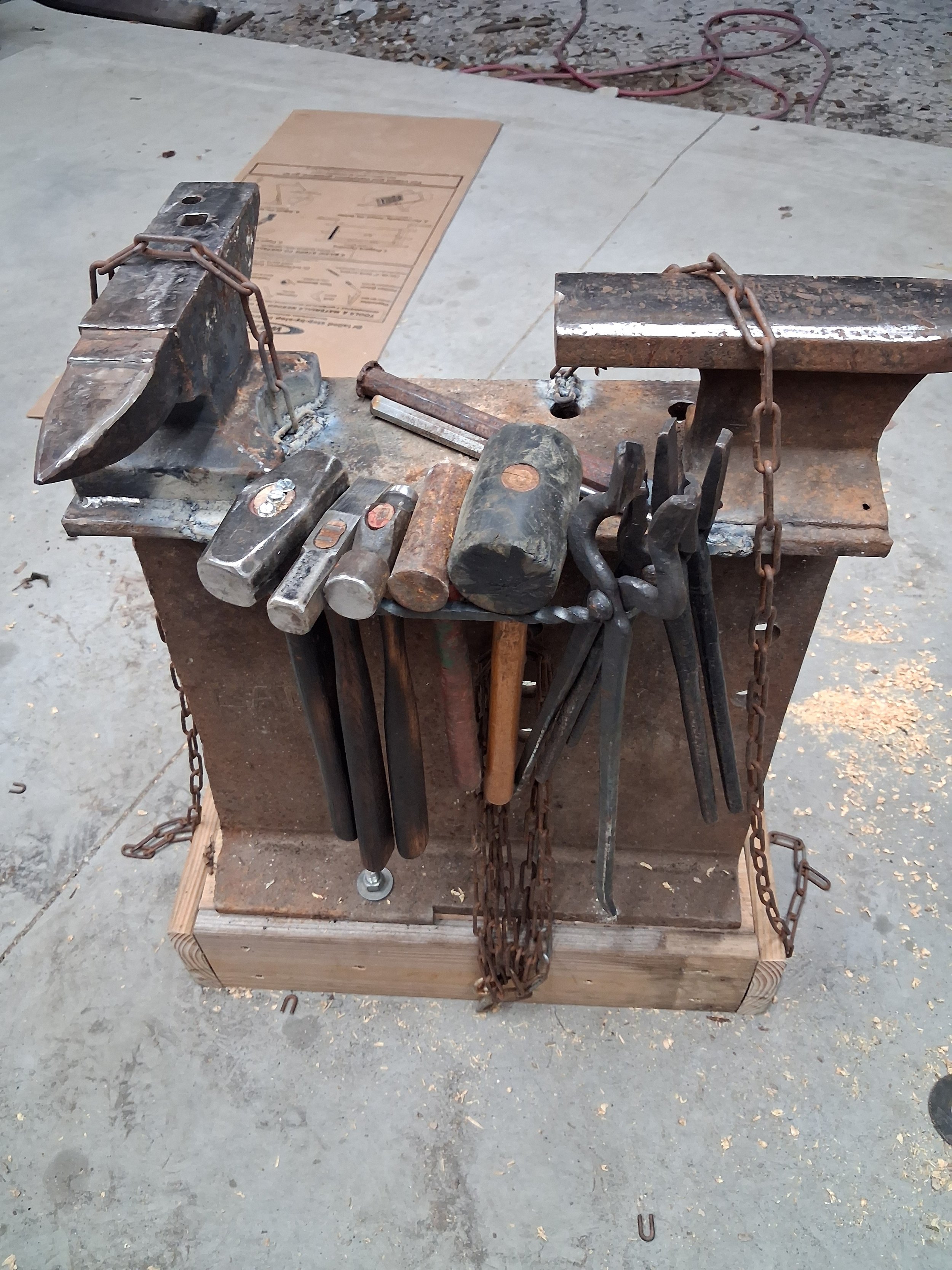 A workbench with various blacksmith tools including hammers, tongs, and chisels, surrounded by chains and set on a concrete floor in a workshop or construction area.