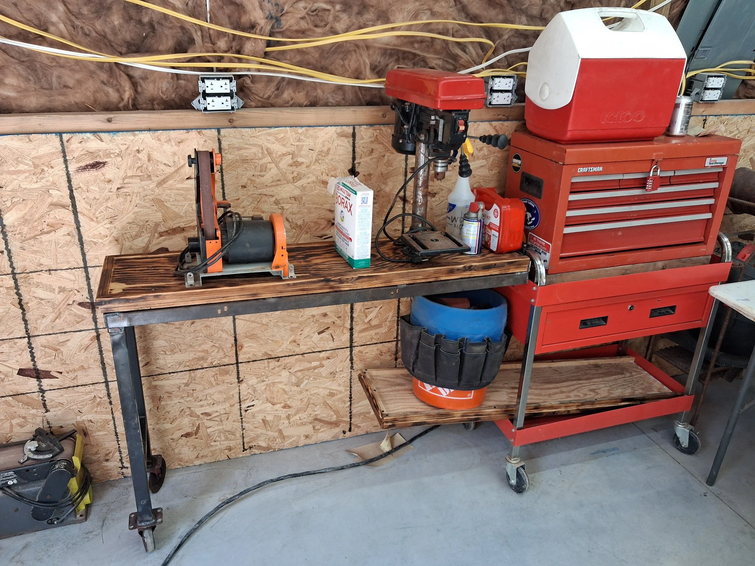 Workshop workspace with tools, electrical outlets on the wall, a red tool chest, and a wooden workbench with various equipment and supplies.