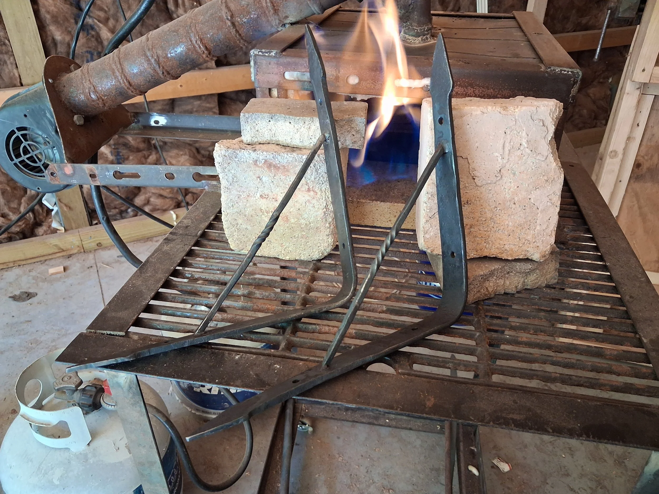 A makeshift outdoor workspace featuring a DIY gas forge with a brick-lined chamber, a propane tank, and a propane hose connected to a blower. The workspace is set in a partially finished wooden structure with insulation in the background.