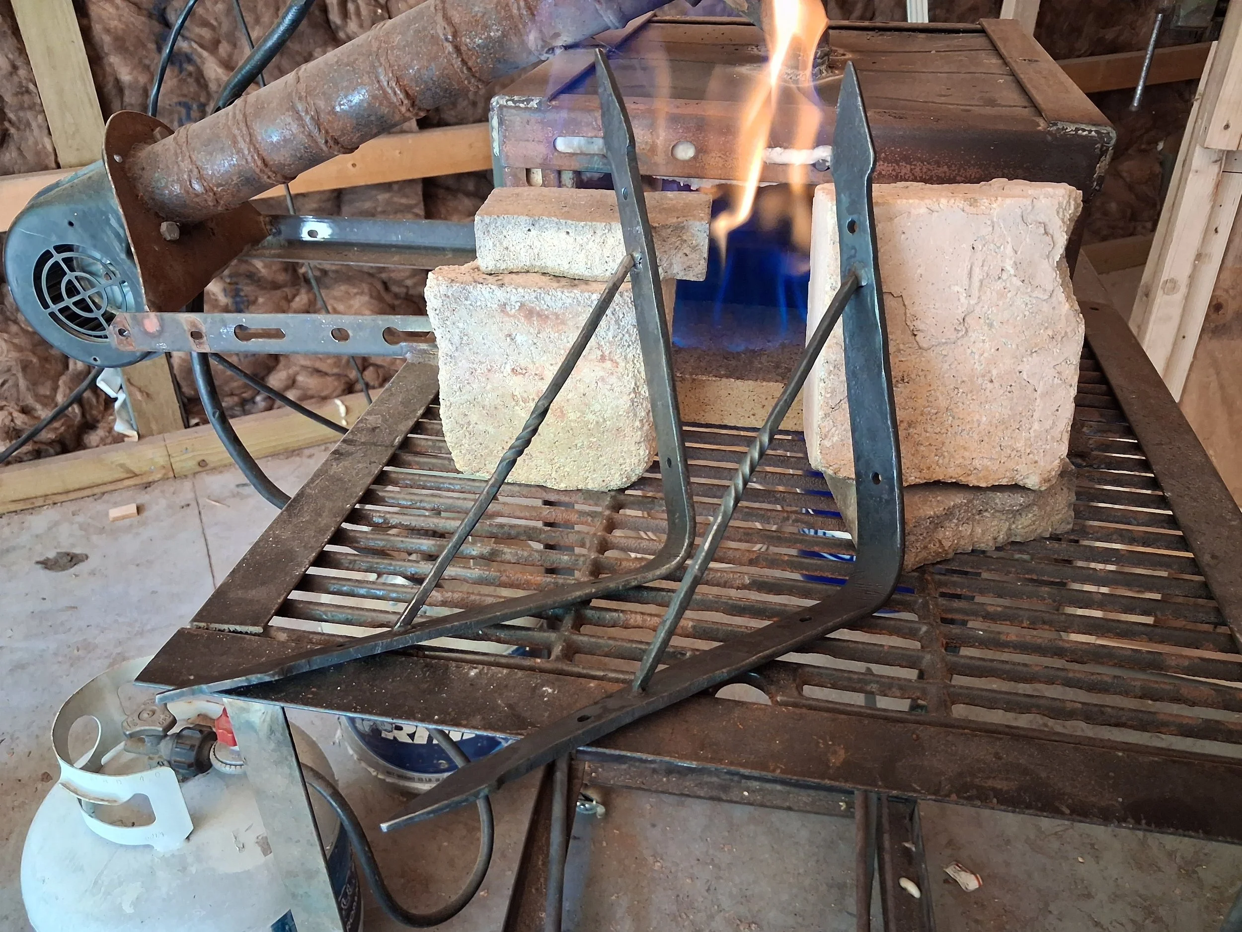 A makeshift outdoor setup with bricks, metal rods, and a propane torch with a flame, possibly for welding or heating metal. The surrounding area appears to be a construction or workshop space with wooden framing and insulation visible in the background.