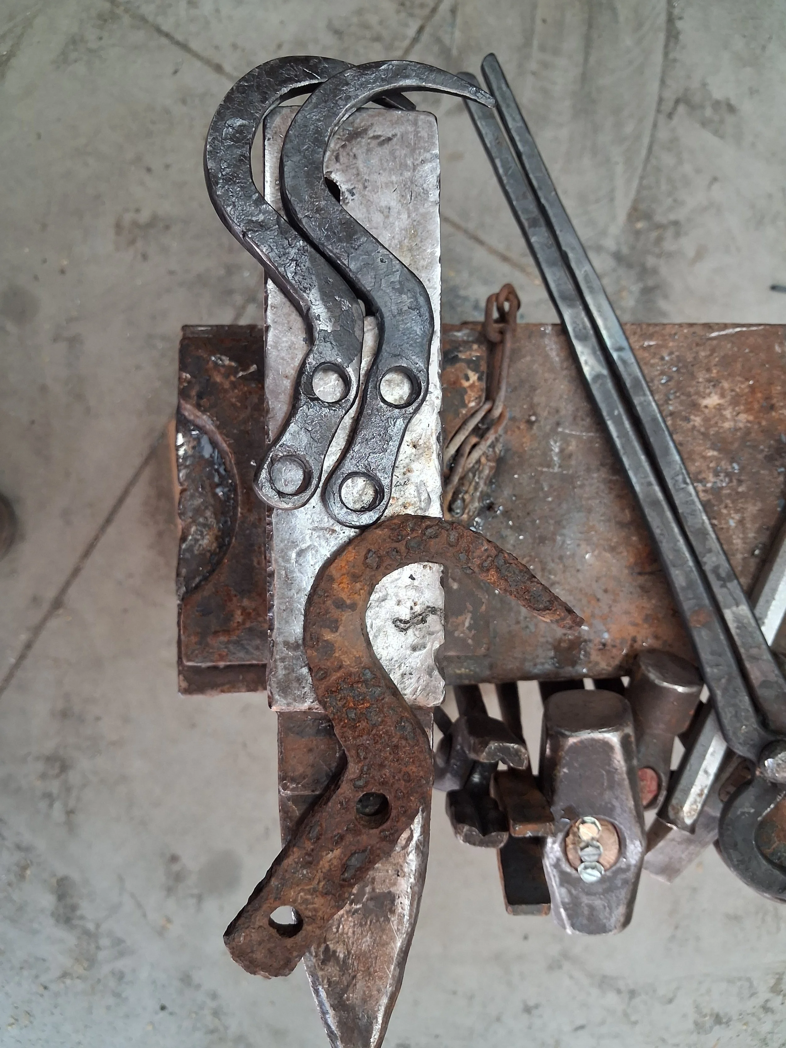 A blacksmith's workspace with curved metal pieces and a chain on a workbench, surrounded by tools and a gray concrete floor.