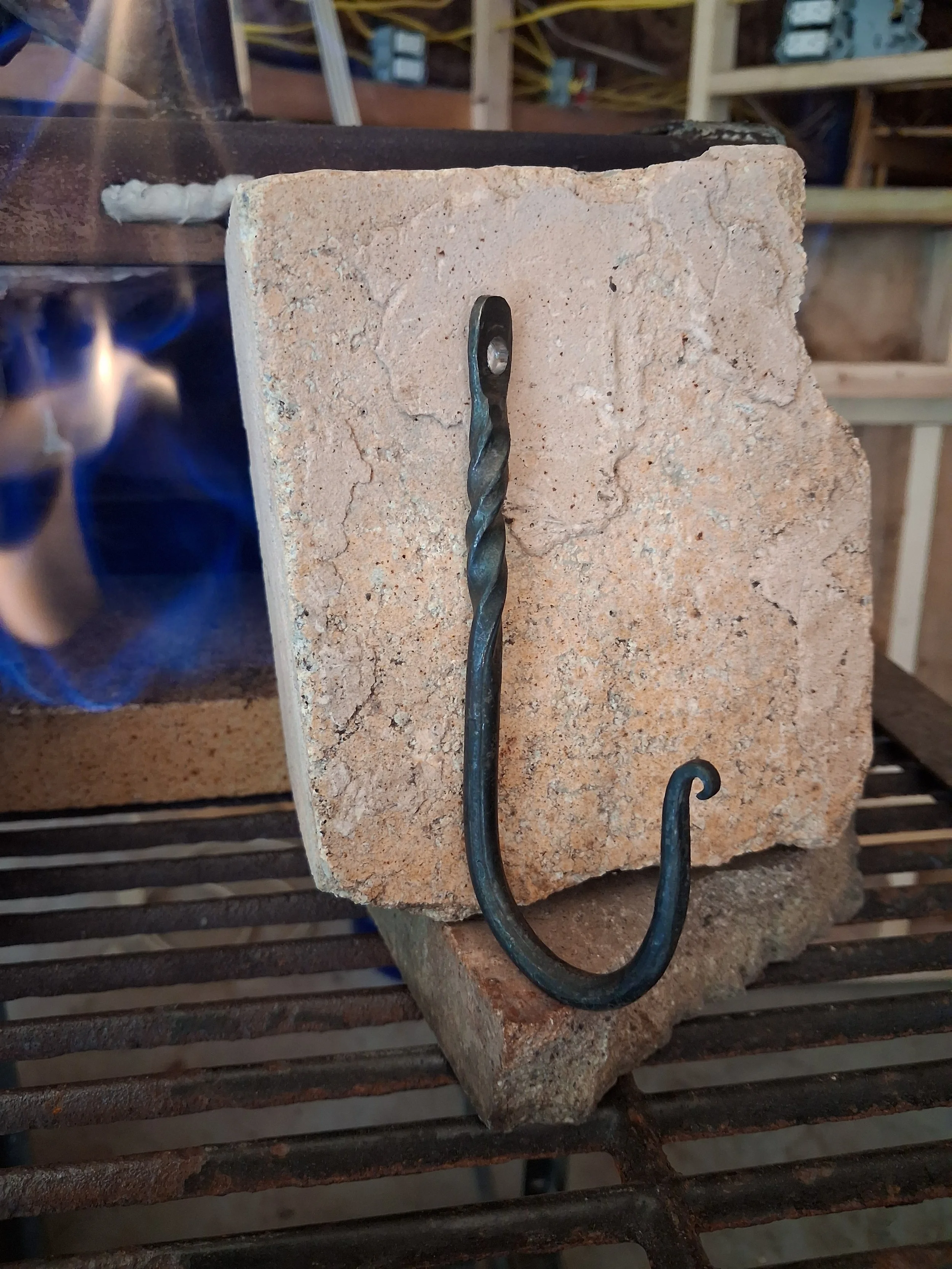 A black, twisted metal hook inserted into a light-colored brick block on a metal grill shelf, with a blue flame visible to the left.