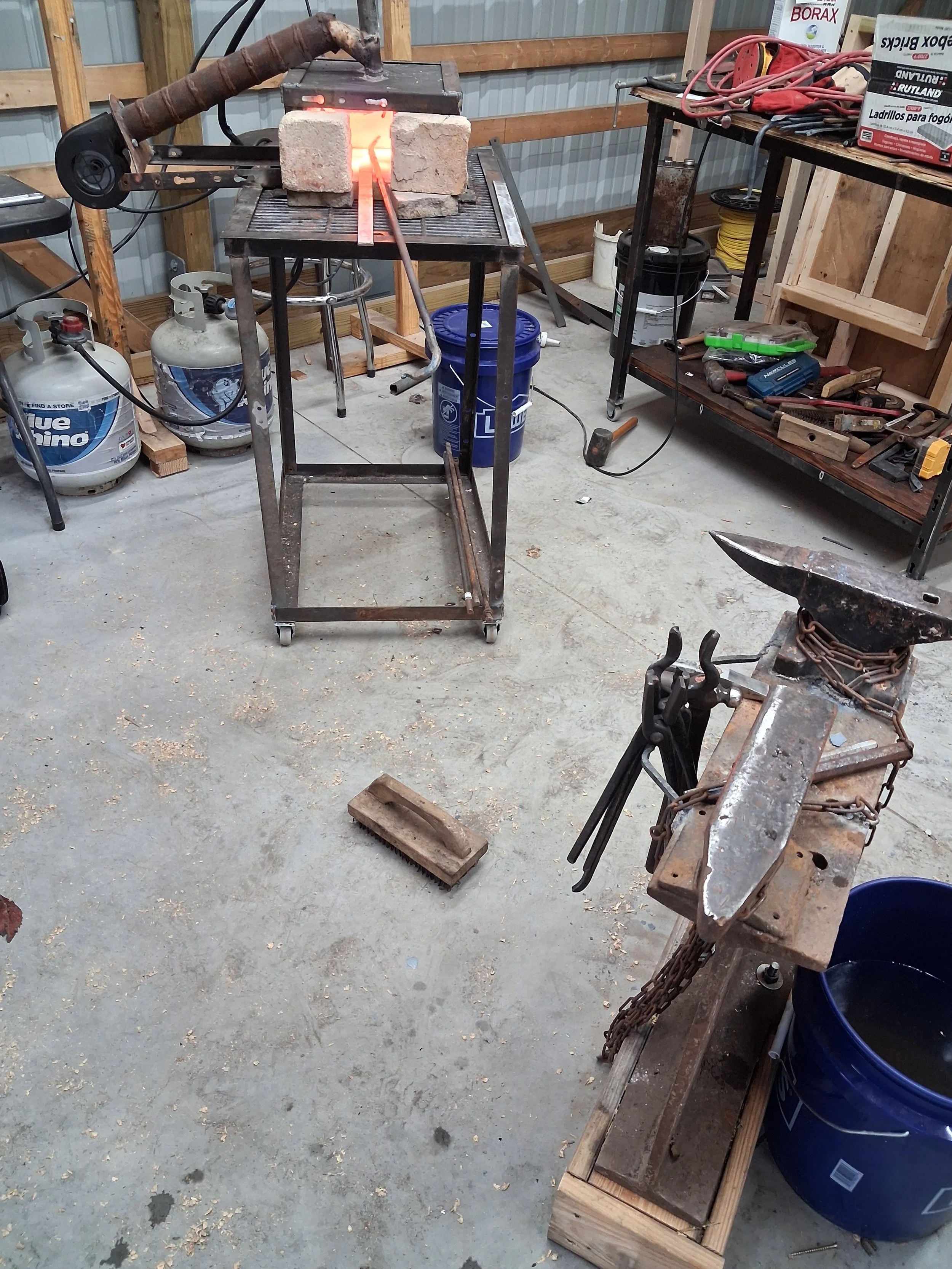 A workbench in a workshop with a forge heated by propane tanks, a black bucket, and various tools and equipment, with metalworking in progress.