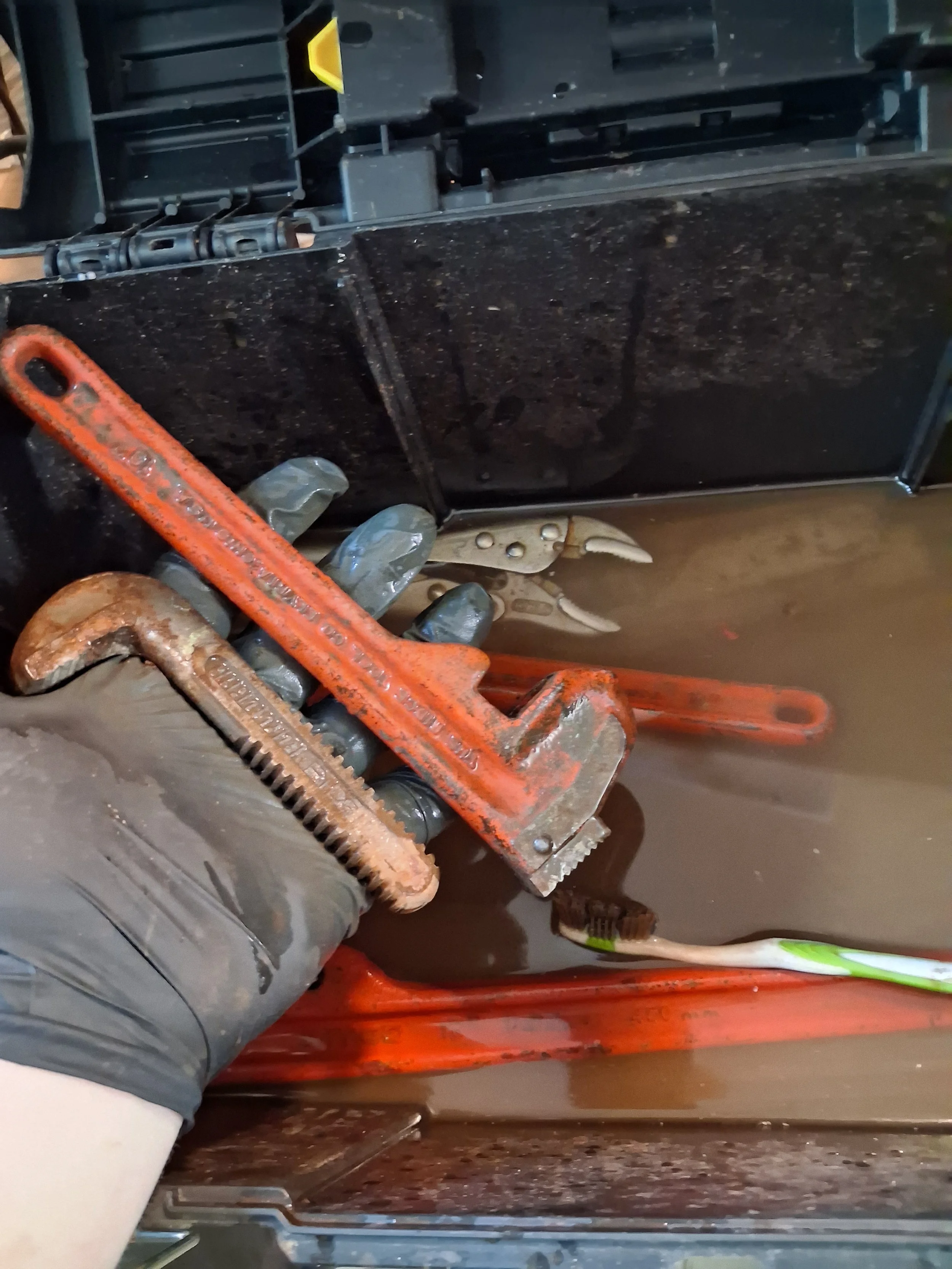 A person's hand wearing a gray glove holding an orange pipe wrench, with a black electrical panel or device and a sickle visible in the background.