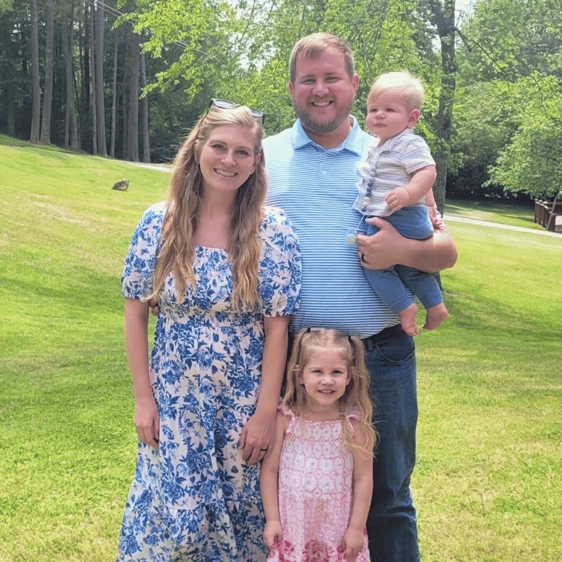 A family of four standing outdoors in a park, smiling for the camera. The mother has long, wavy blonde hair and is wearing a blue and white floral dress. The father has a beard, short hair, and is wearing a light blue striped shirt, holding a young boy in his arm. The young boy has blonde hair and is wearing a striped shirt and dark pants. The daughter has curly blonde hair and is dressed in a pink patterned dress, standing in front of her mother.