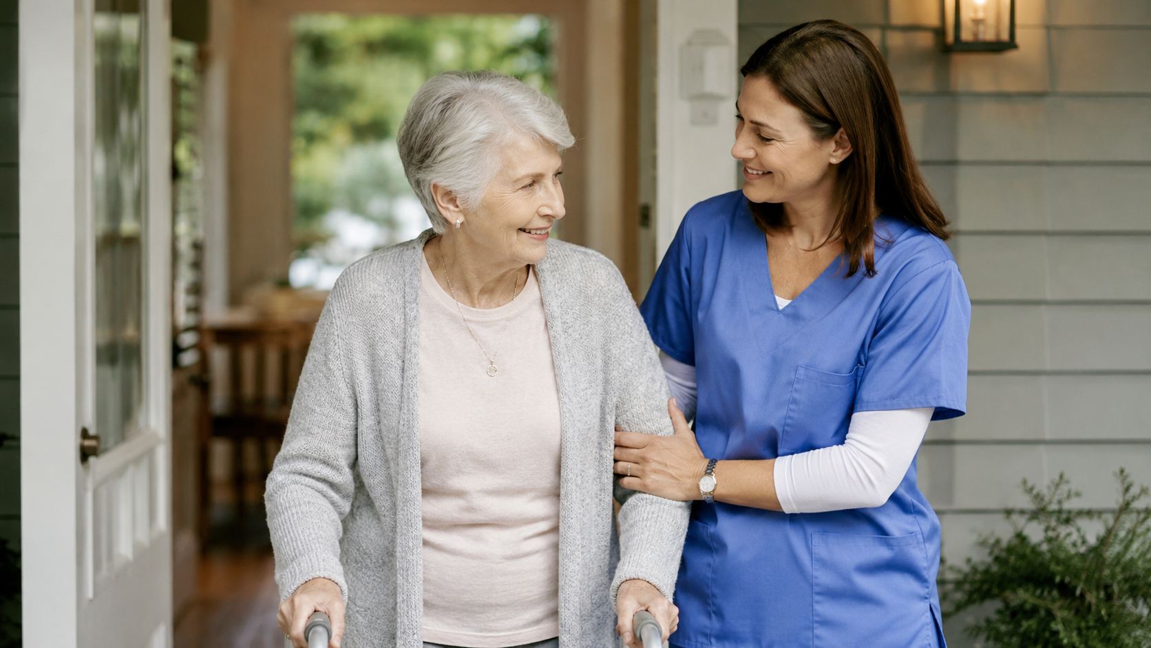 A healthcare worker, wearing blue scrubs, is assisting an elderly woman using a walker outside her house.