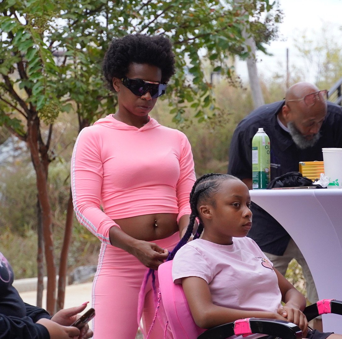 A young girl sits in a pink wheelchair while a woman in pink workout clothes styles her hair. The woman has dark sunglasses and a belly button ring. An older man is in the background, leaning over a table with various items on it, including a spray c