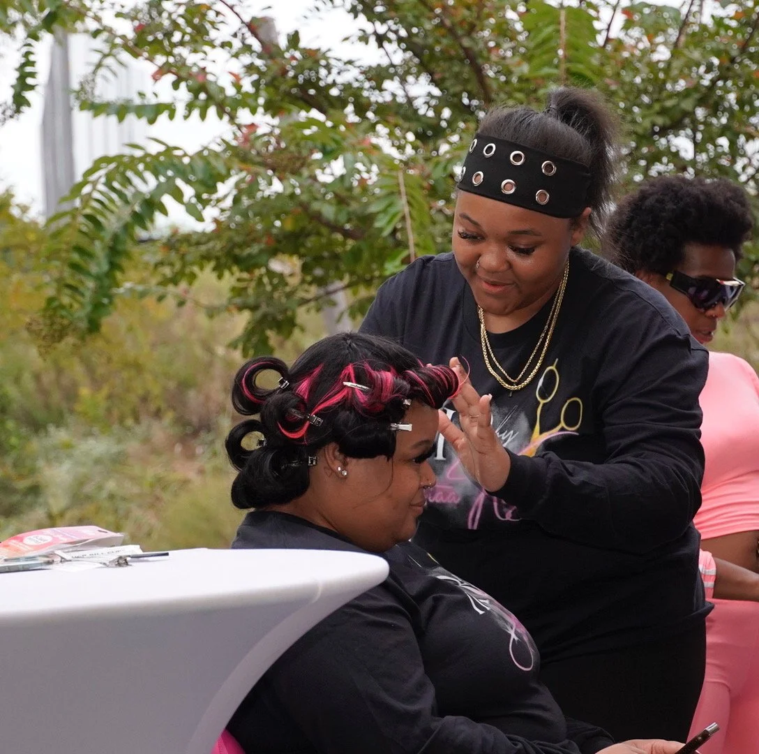 A woman with pink and black curlers in her hair getting her makeup done outdoors, smiling as another woman styles her eyebrows. Another woman in pink stands nearby.